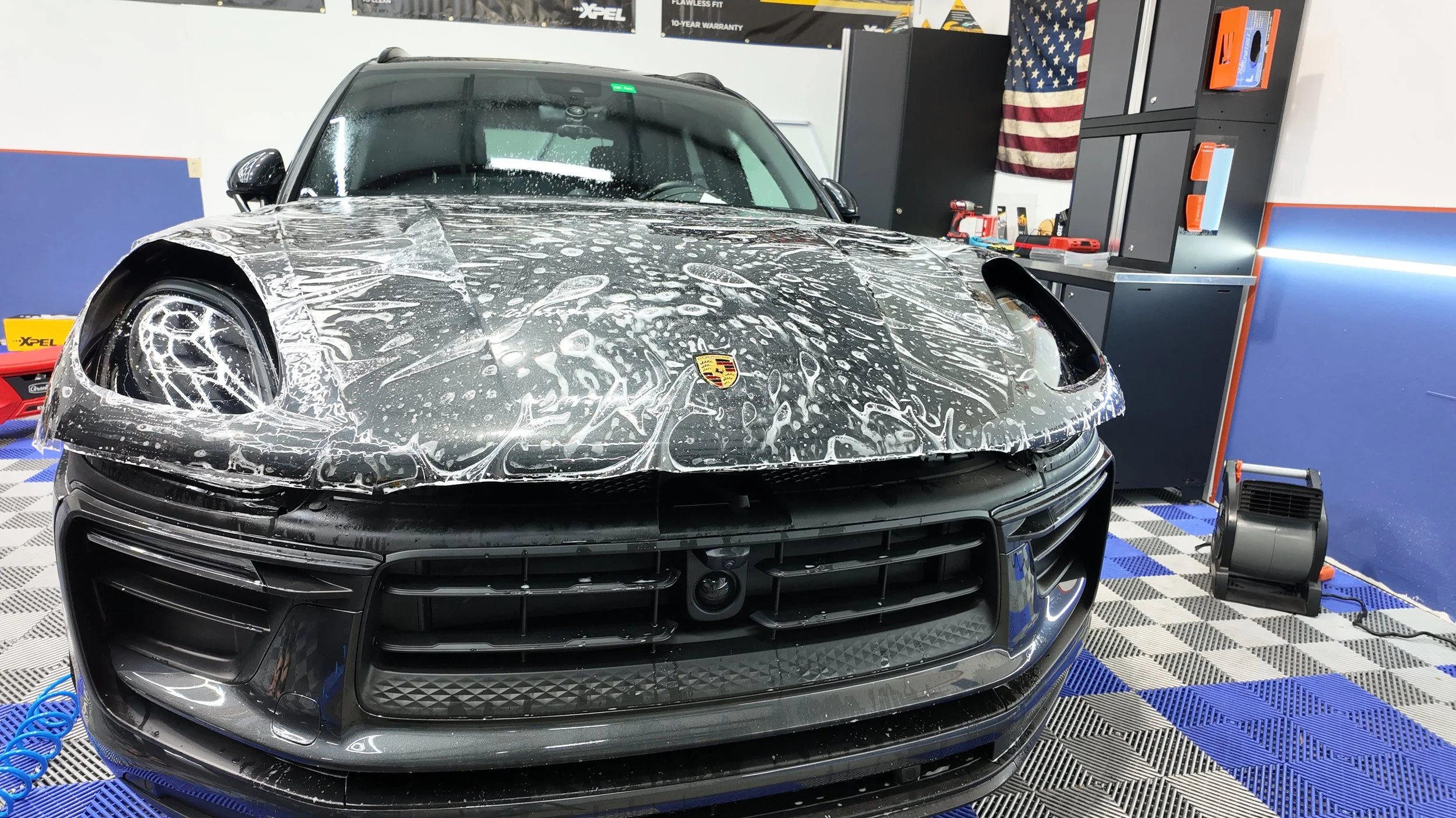 A black Porsche Macan undergoing a car wash or detail, with soap suds on its hood and front, in a garage or detailing shop.