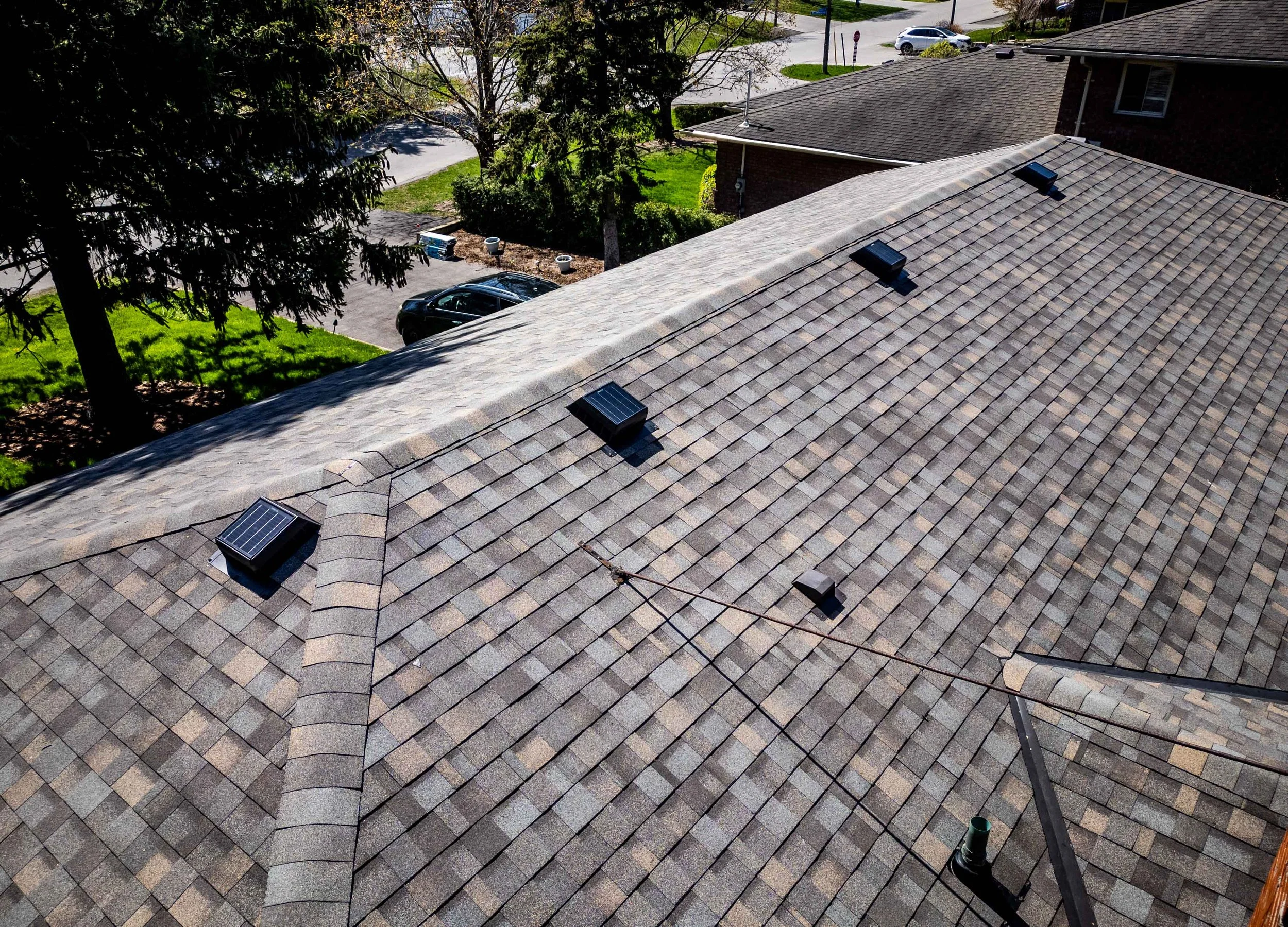 Overhead view of a residential roof with asphalt shingles, showing solar vents installed, surrounded by greenery in a suburban area.