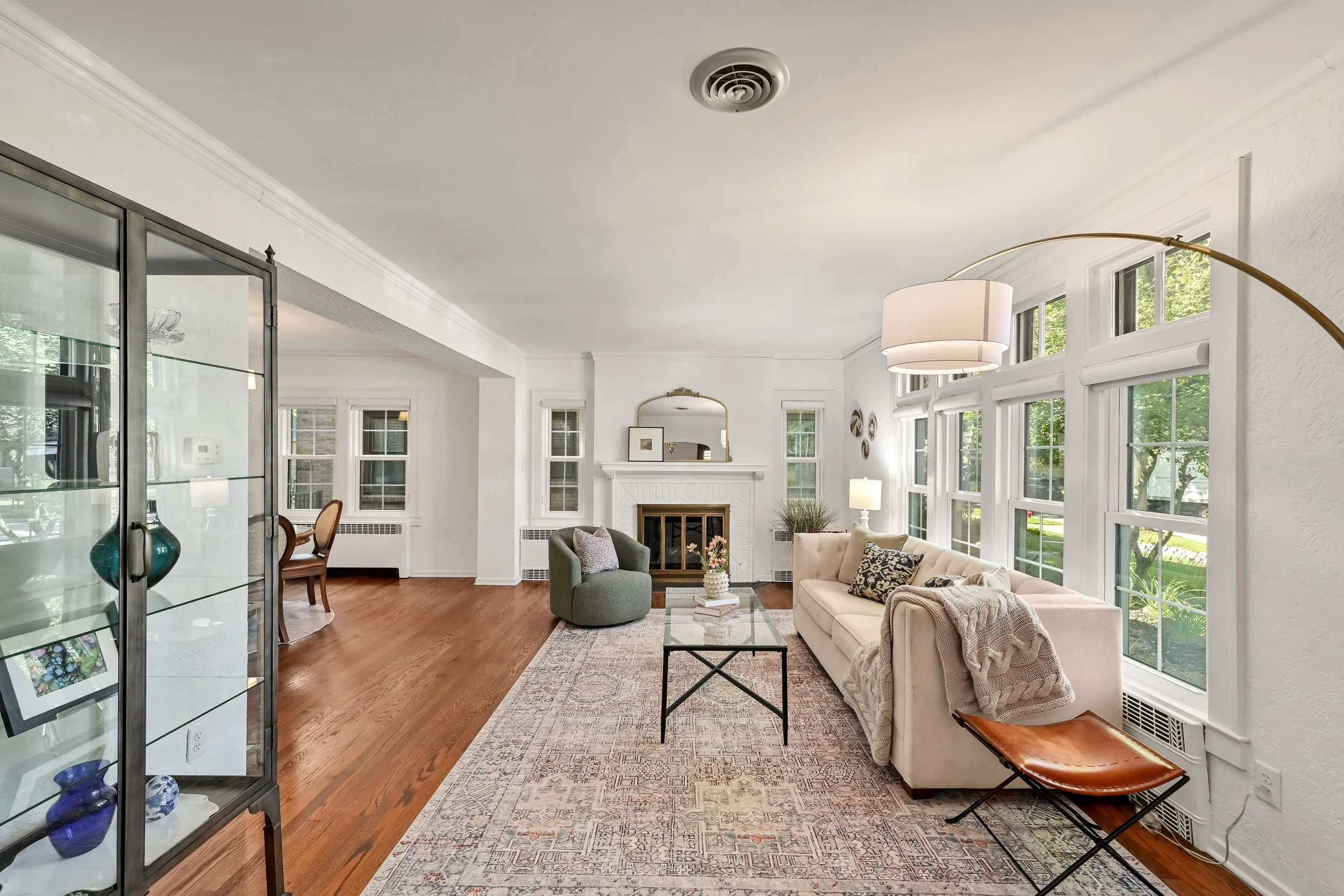Bright living room with white walls, large windows, a gas fireplace, beige sofa, gray armchair, glass coffee table, patterned area rug, and decorative items including a mirror, plants, and vases.