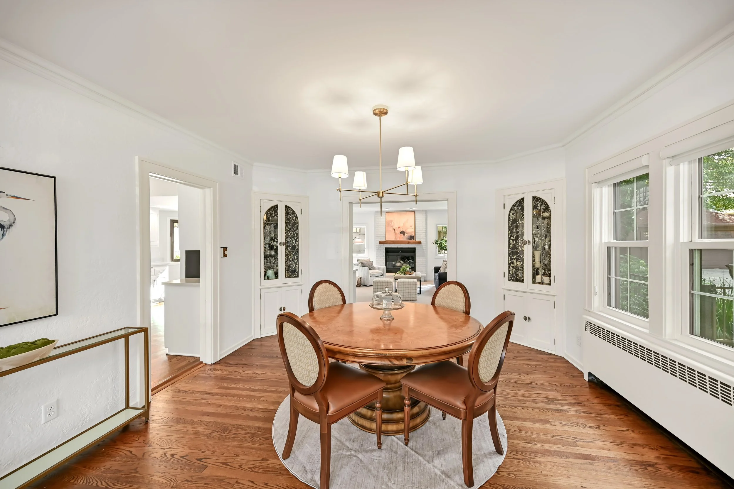 A dining room with a oval wooden table and six chairs, a chandelier with six white shades, white cabinets with glass doors, large windows, and a view of a living room with a fireplace across the room.