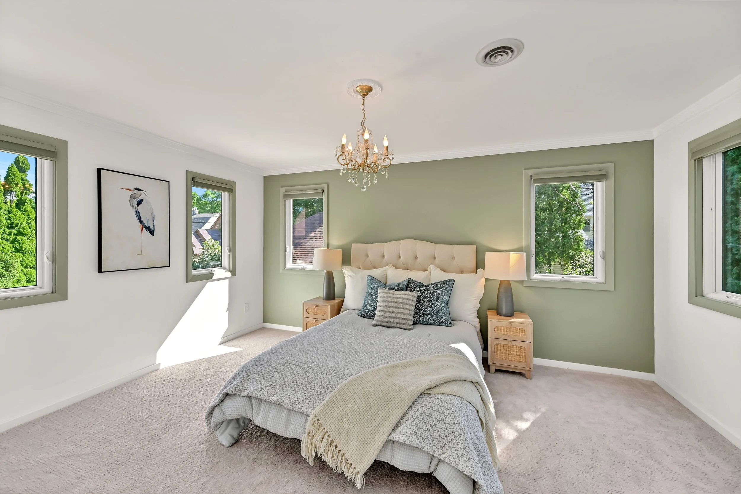 A bedroom with a bed, two nightstands with lamps, four windows showing trees outside, a painting of a heron on the wall, and a chandelier hanging from the ceiling.