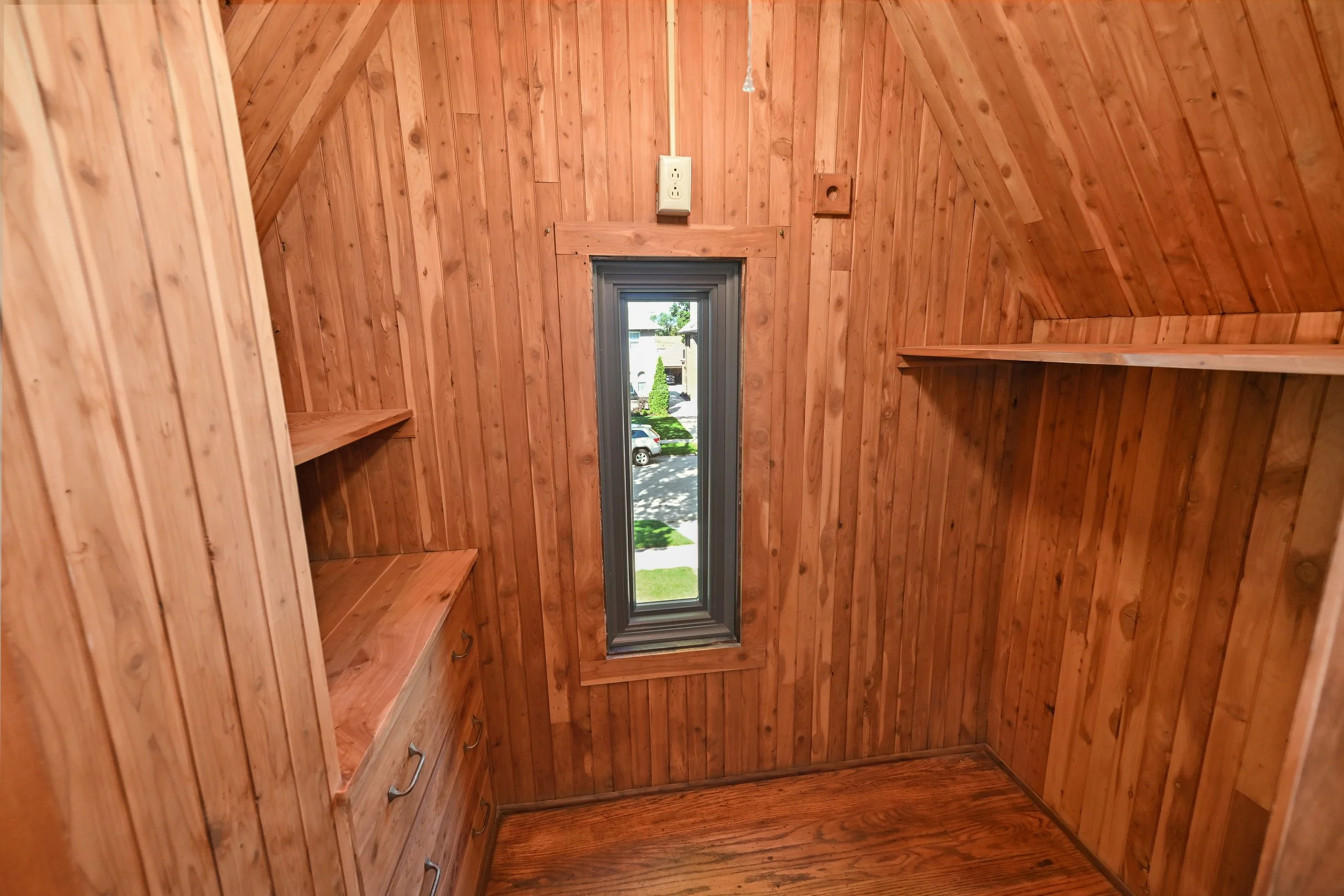 Inside a small wooden room with angled walls, featuring built-in wooden shelves, a window with a view of parked cars and trees outside, and an electrical outlet near the ceiling.