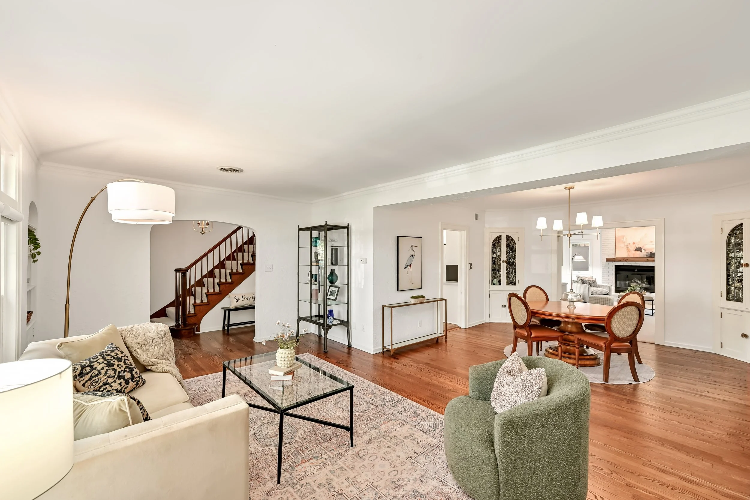 Living room with hardwood floors, beige sofa and green armchair, glass coffee table, area rug, white walls, staircase, and dining area visible in the background.