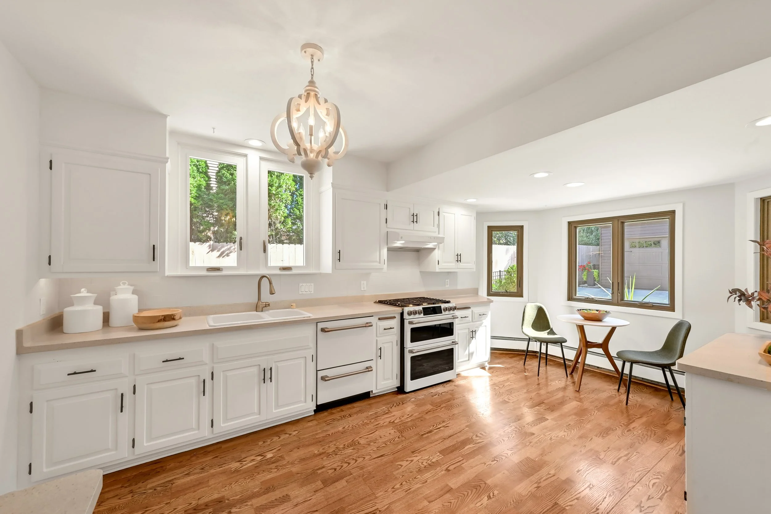 Bright kitchen with white cabinets, beige countertops, a stove, three windows, and a small dining area with two chairs and a round table.