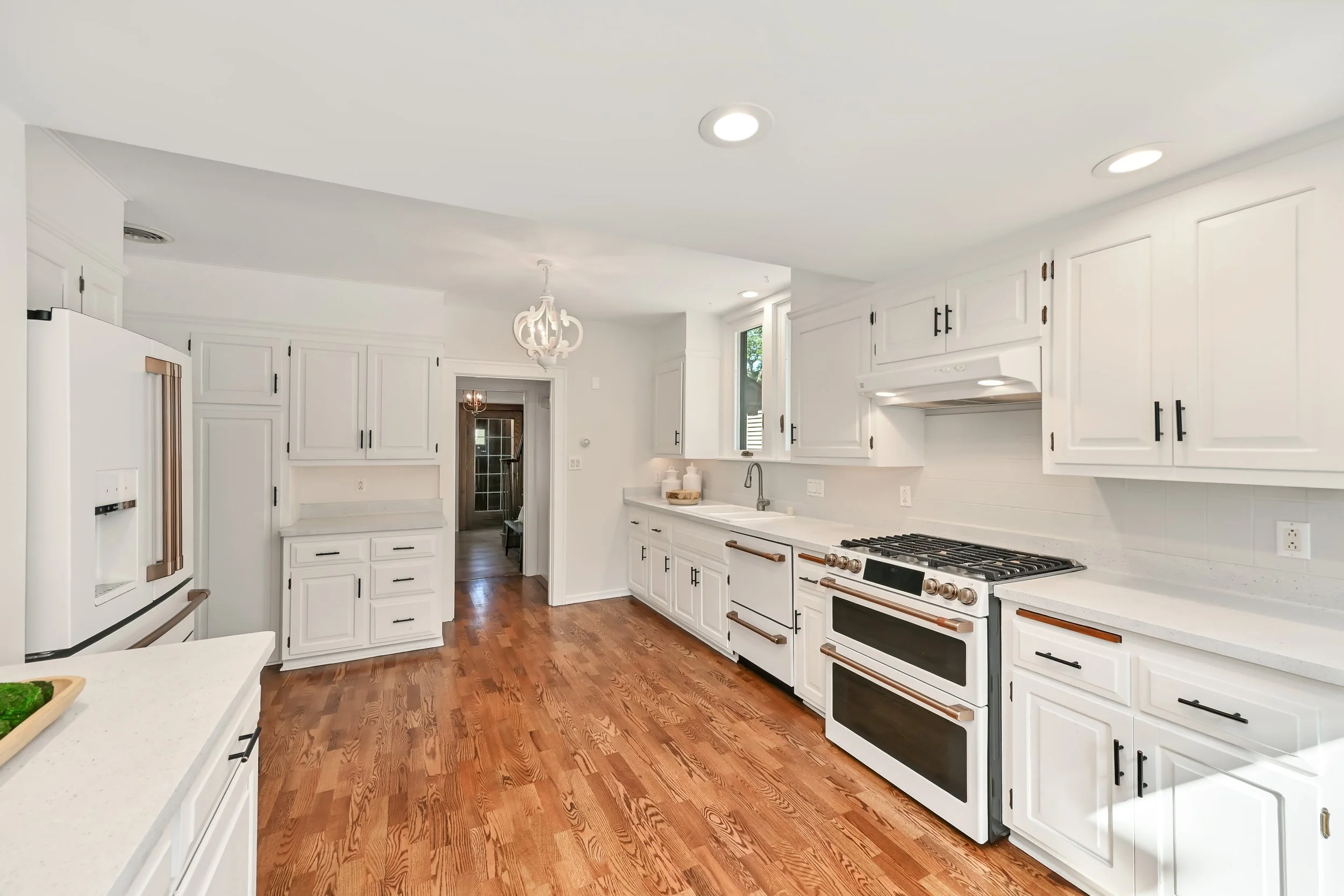Modern white kitchen with hardwood floors, white cabinets, stainless steel appliances, and a gas stove. A small window above the sink lets in natural light, and there is a doorway leading to another room.