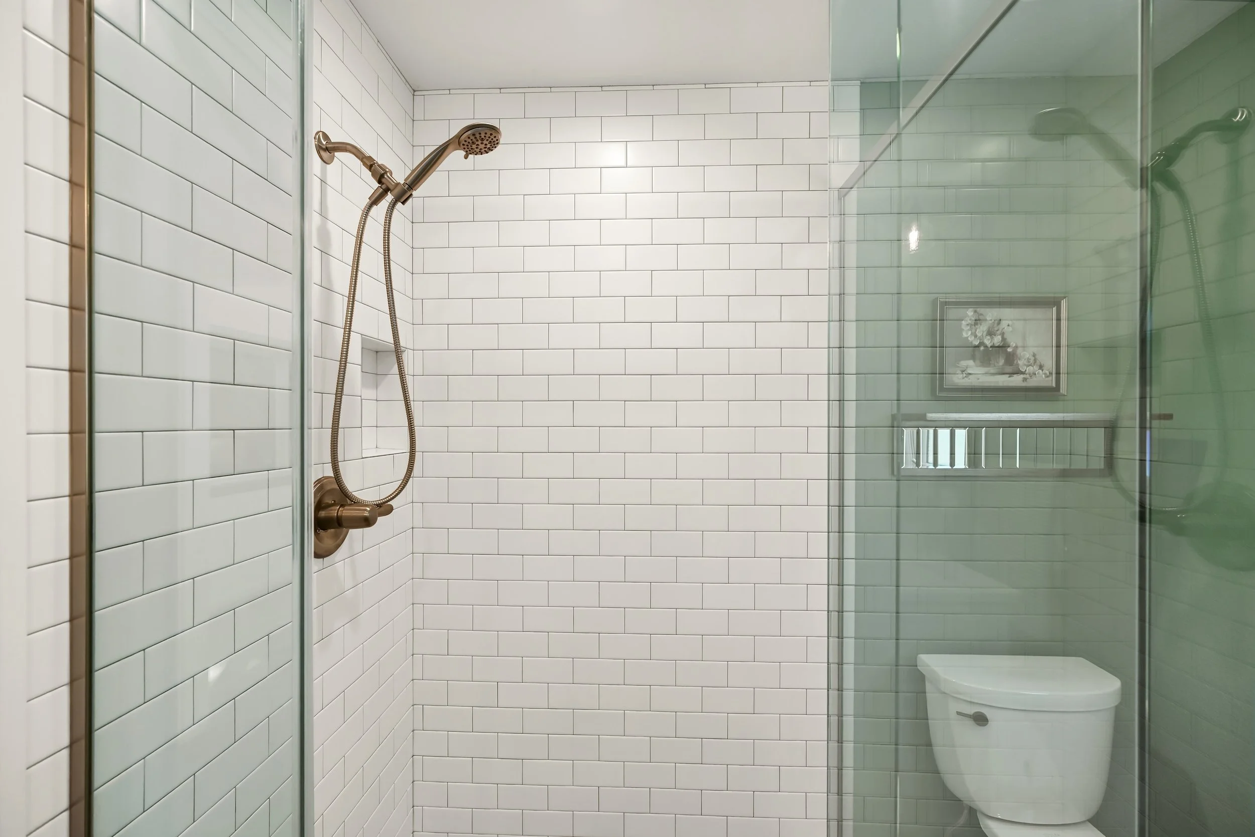 A modern bathroom shower with white subway tiles, a bronze showerhead, and a glass enclosure. A framed picture and a toilet are visible outside the shower.