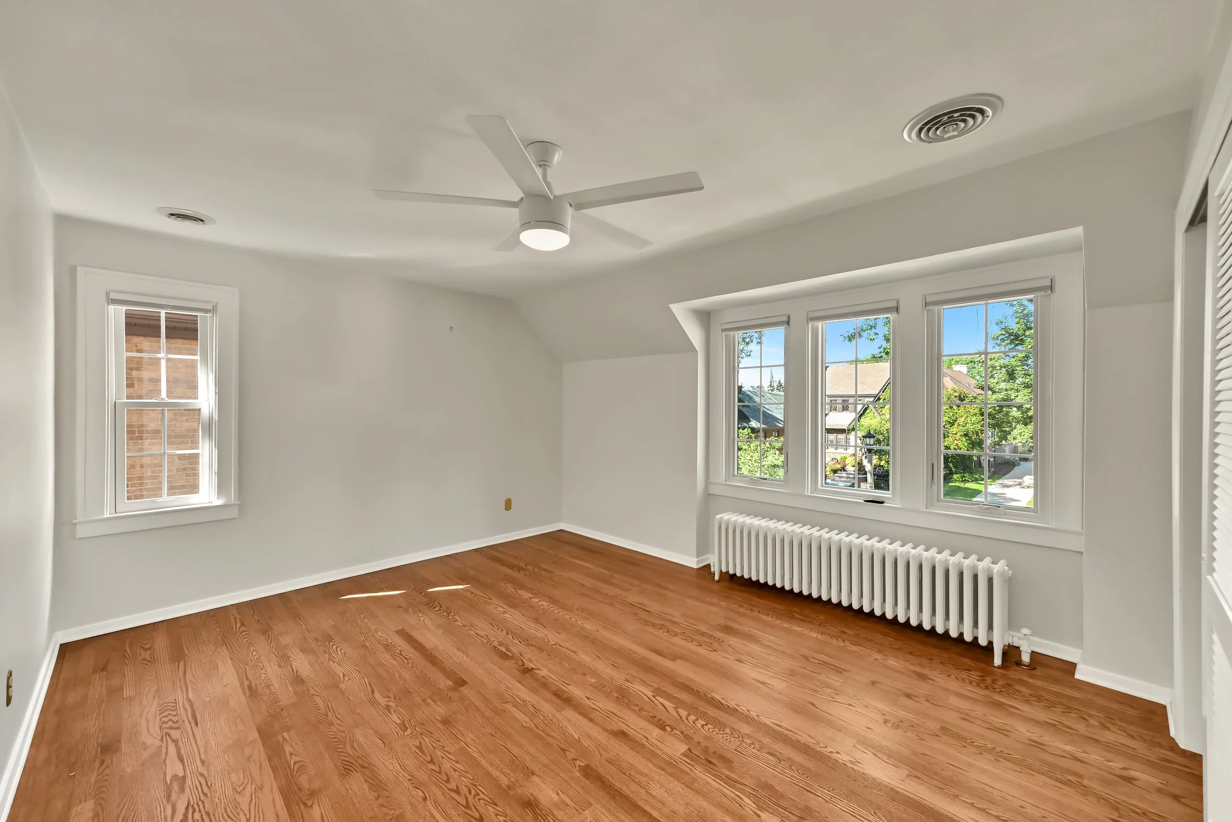 Empty room with white walls, hardwood floors, two windows with white trims and view of trees and houses outside, ceiling fan with light, radiator near window, and ceiling vents.