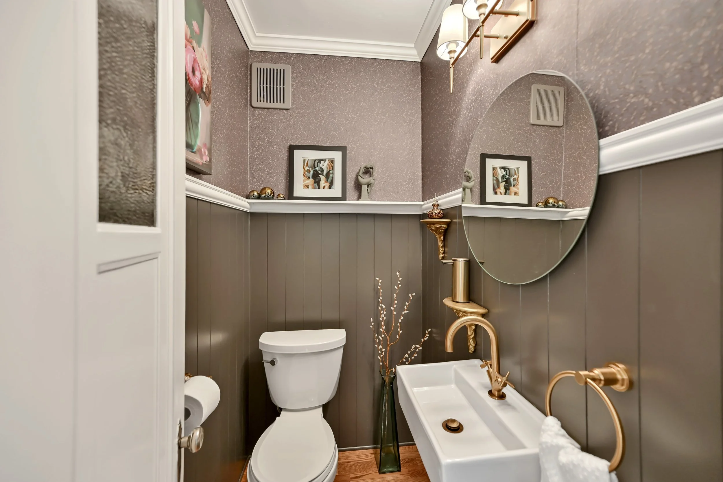 A small bathroom with a white toilet, dark brown beadboard wainscoting, a white sink with gold fixtures, and framed artwork on the wall. The mirror reflects the wall and artwork.
