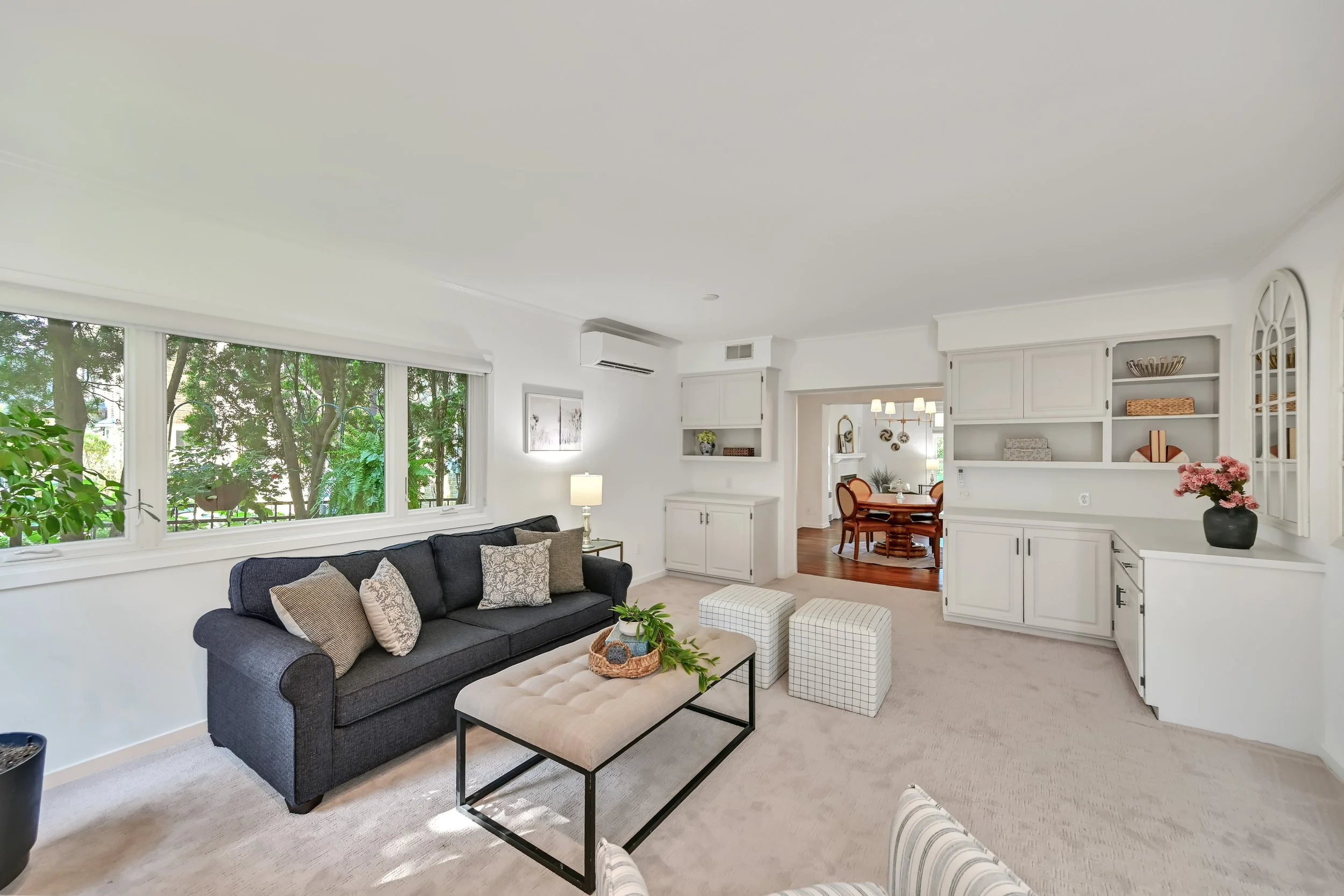 Living room with large front-facing window, dark gray sofa with cushions, beige ottoman, two white checkered stools, plants, and a view into a dining area with a round table and chairs, white built-in shelves, and decor.