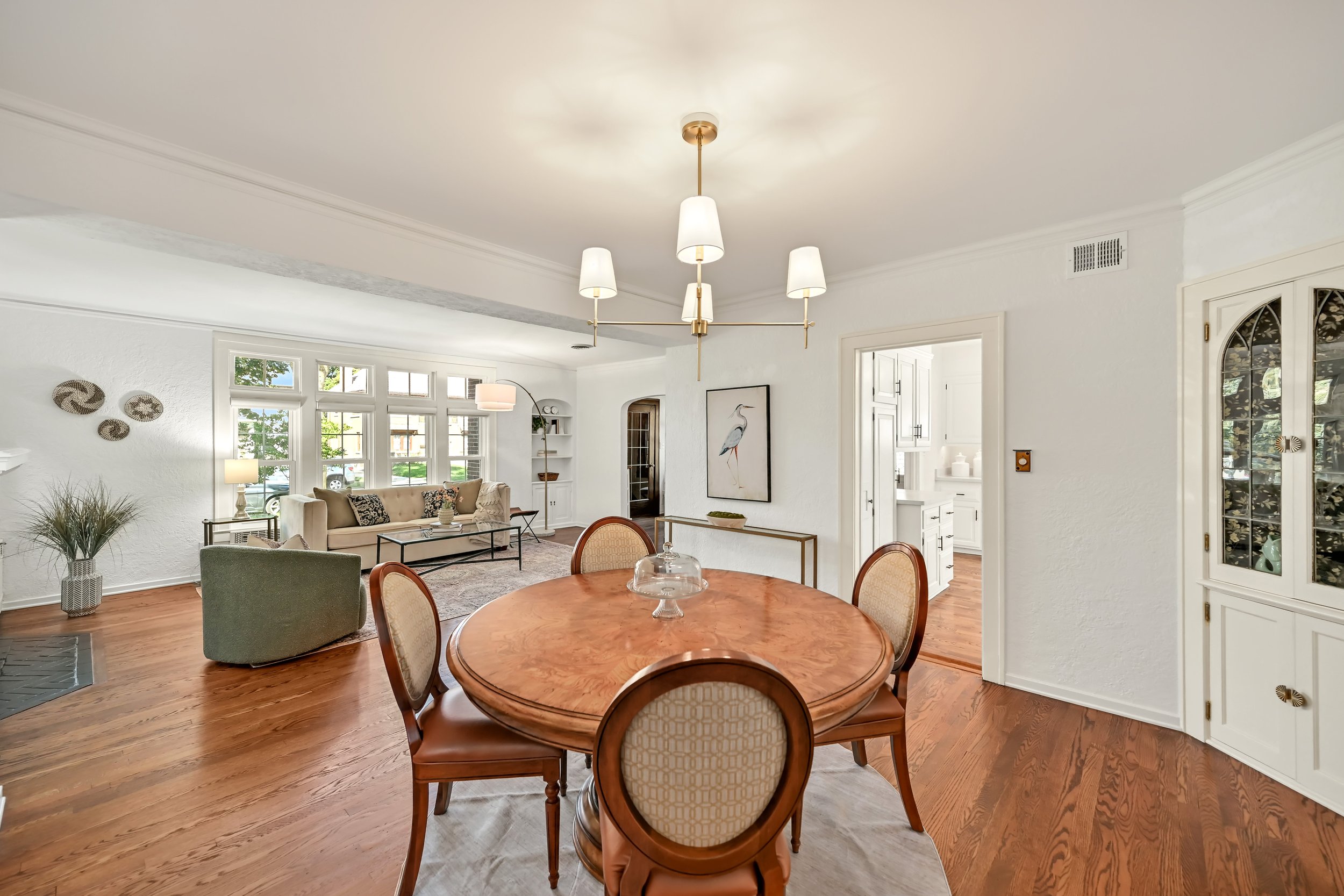 Living room and dining area with white walls, wooden floors, a round wooden table with chairs, a chandelier, a beige sofa, a green armchair, a painting of a heron, and large windows providing natural light.