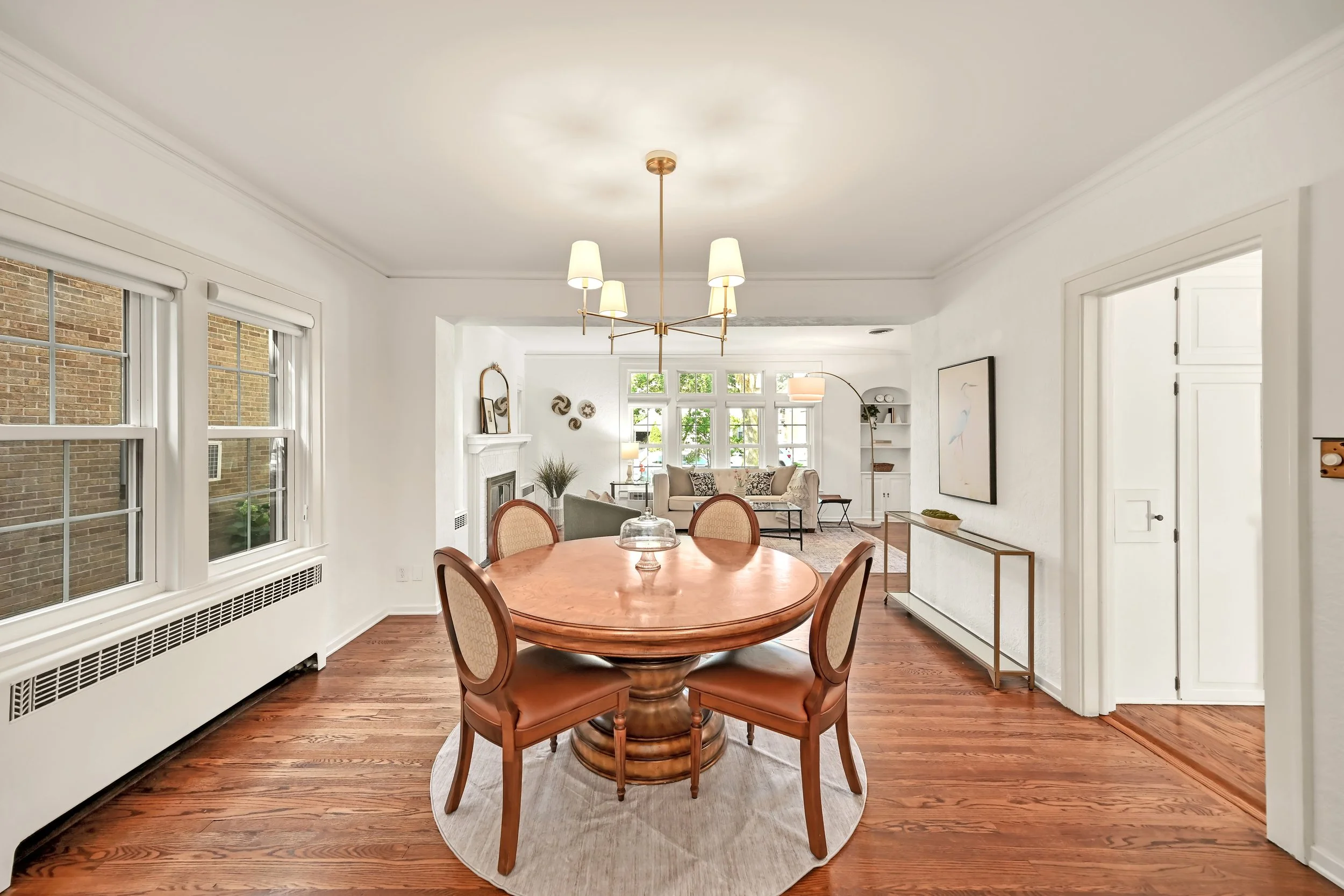 A dining area with a round wooden table and four matching chairs, situated on a circular rug. The room features large windows with white trim, white walls, and hardwood floors. Modern light fixtures and decorative wall art are visible.