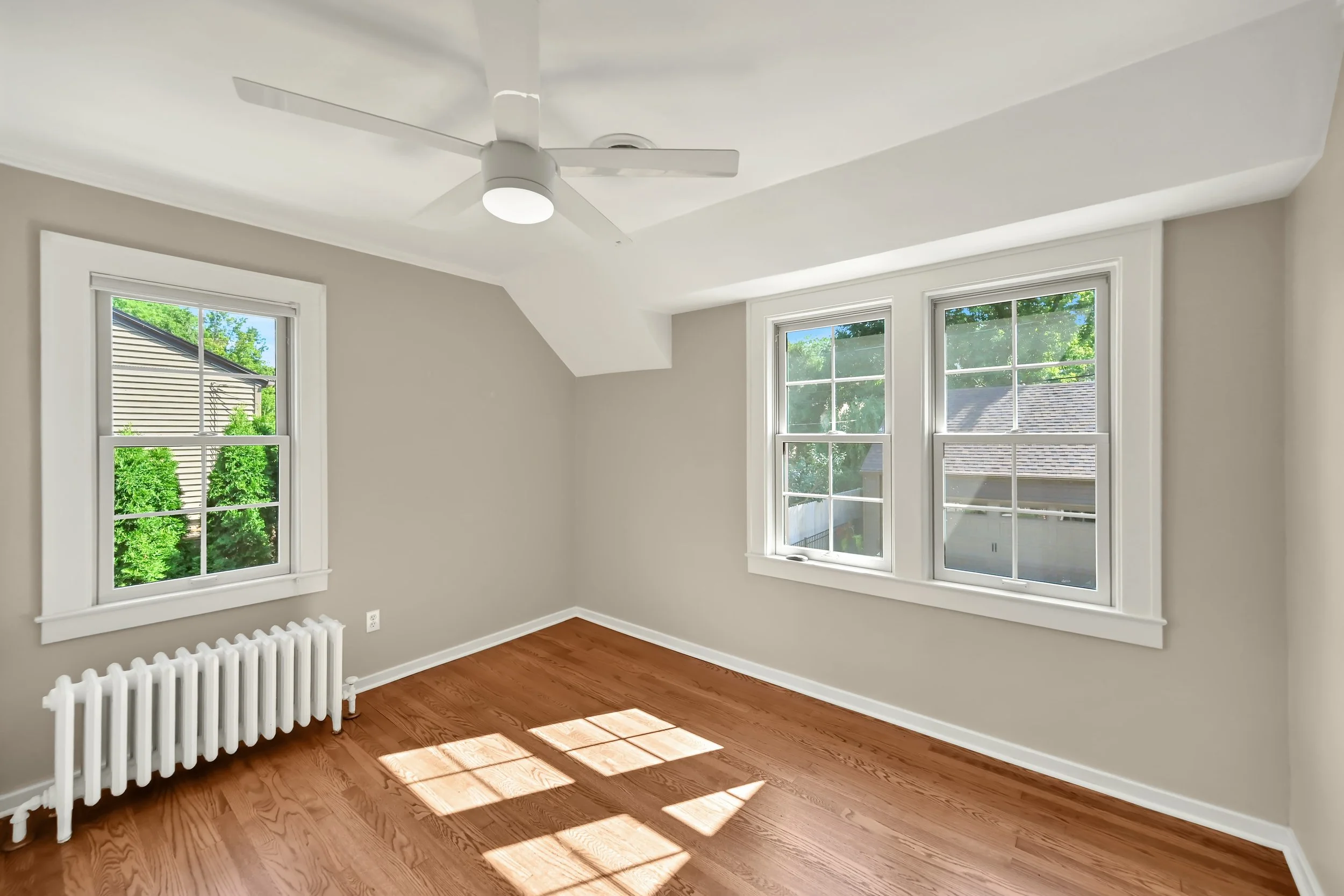 Empty room with hardwood floors, white walls, two large double-hung windows, a white ceiling fan, and a white radiator, with sunlight casting shadows on the floor.
