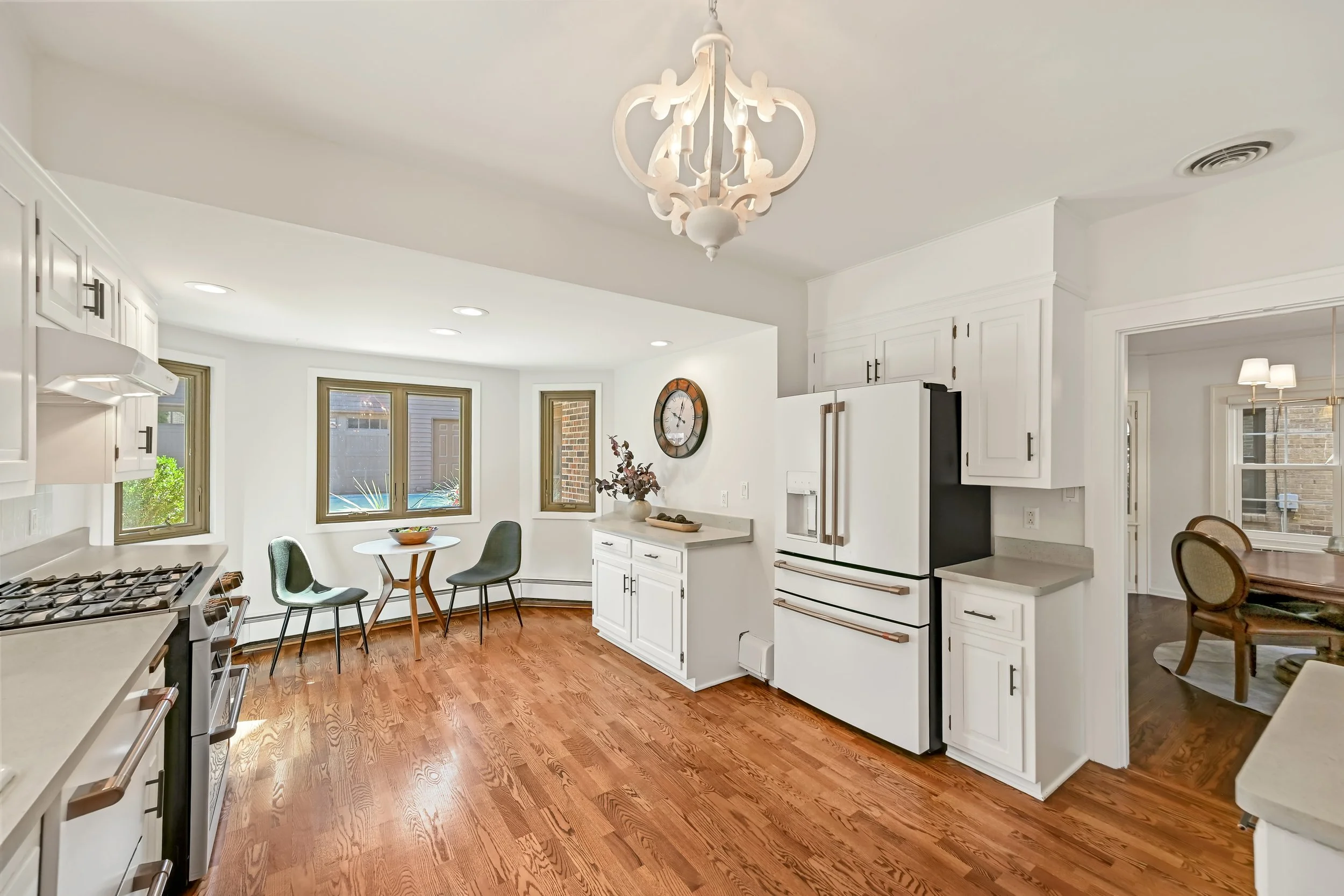 Bright kitchen with white cabinets, wooden floors, a round table with chairs, a wall clock, and large windows allowing natural light.