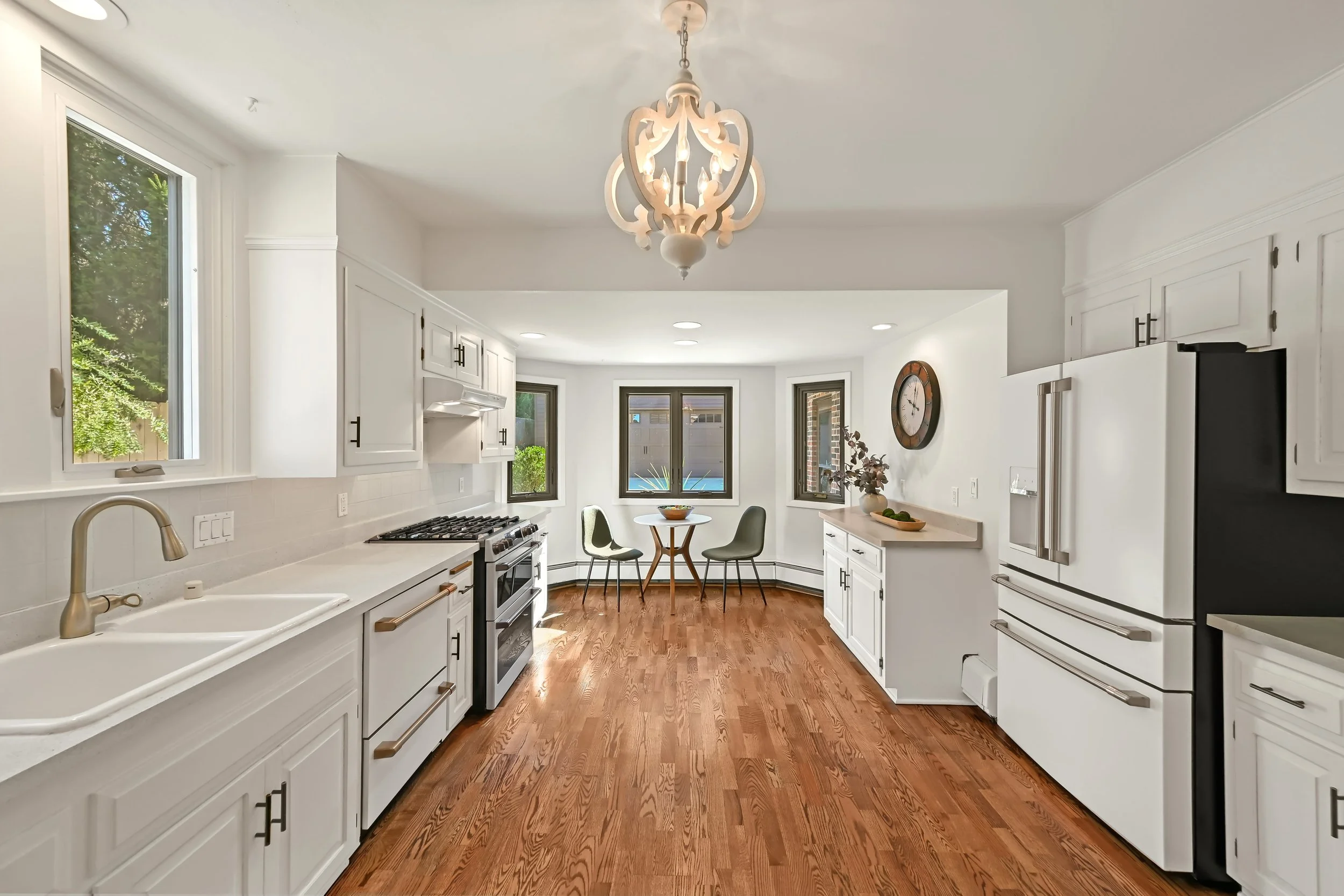 A bright white kitchen with hardwood floors, large windows, white cabinets, and stainless steel appliances. There is a small round dining table with three chairs near the window.