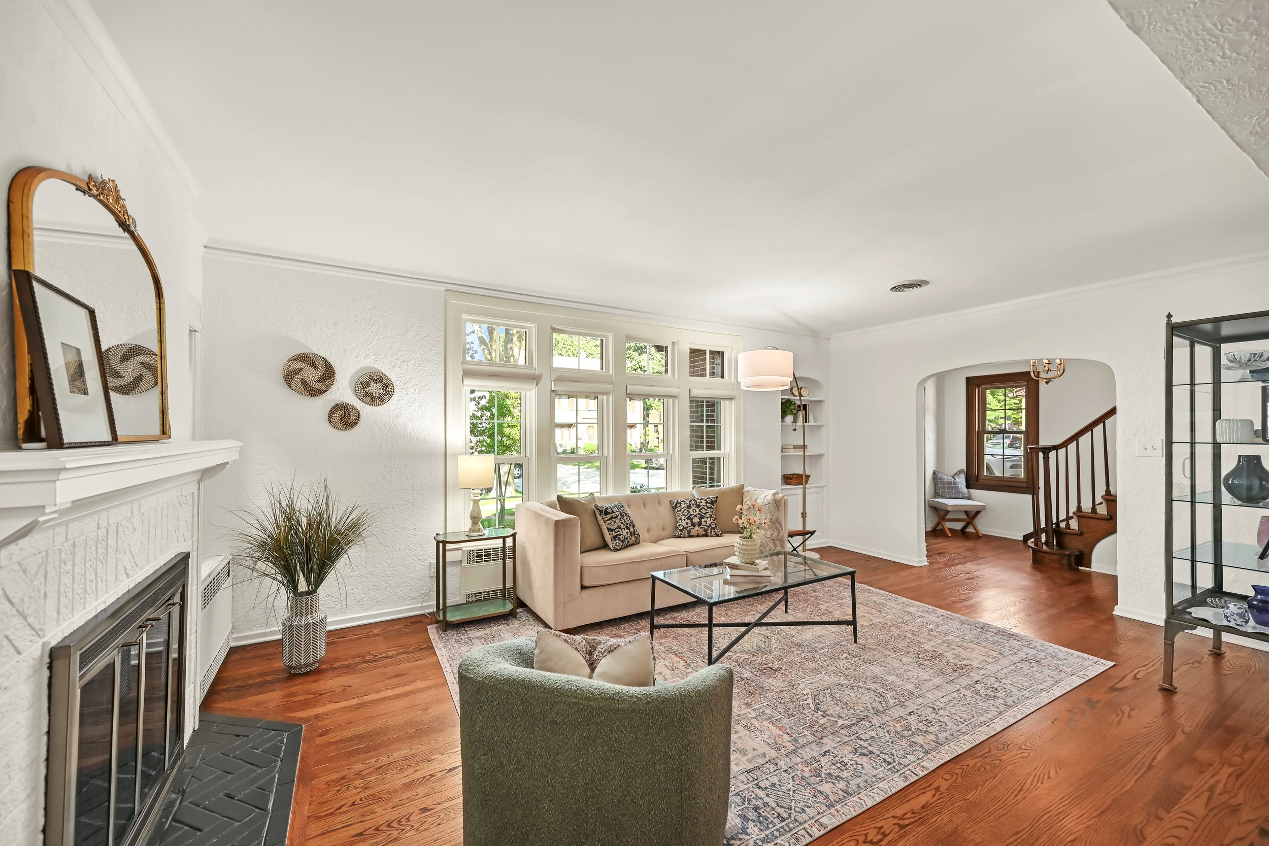 Living room with white walls, large windows, beige sofa, glass coffee table, area rug, fireplace, bookshelf, and various decorative items.