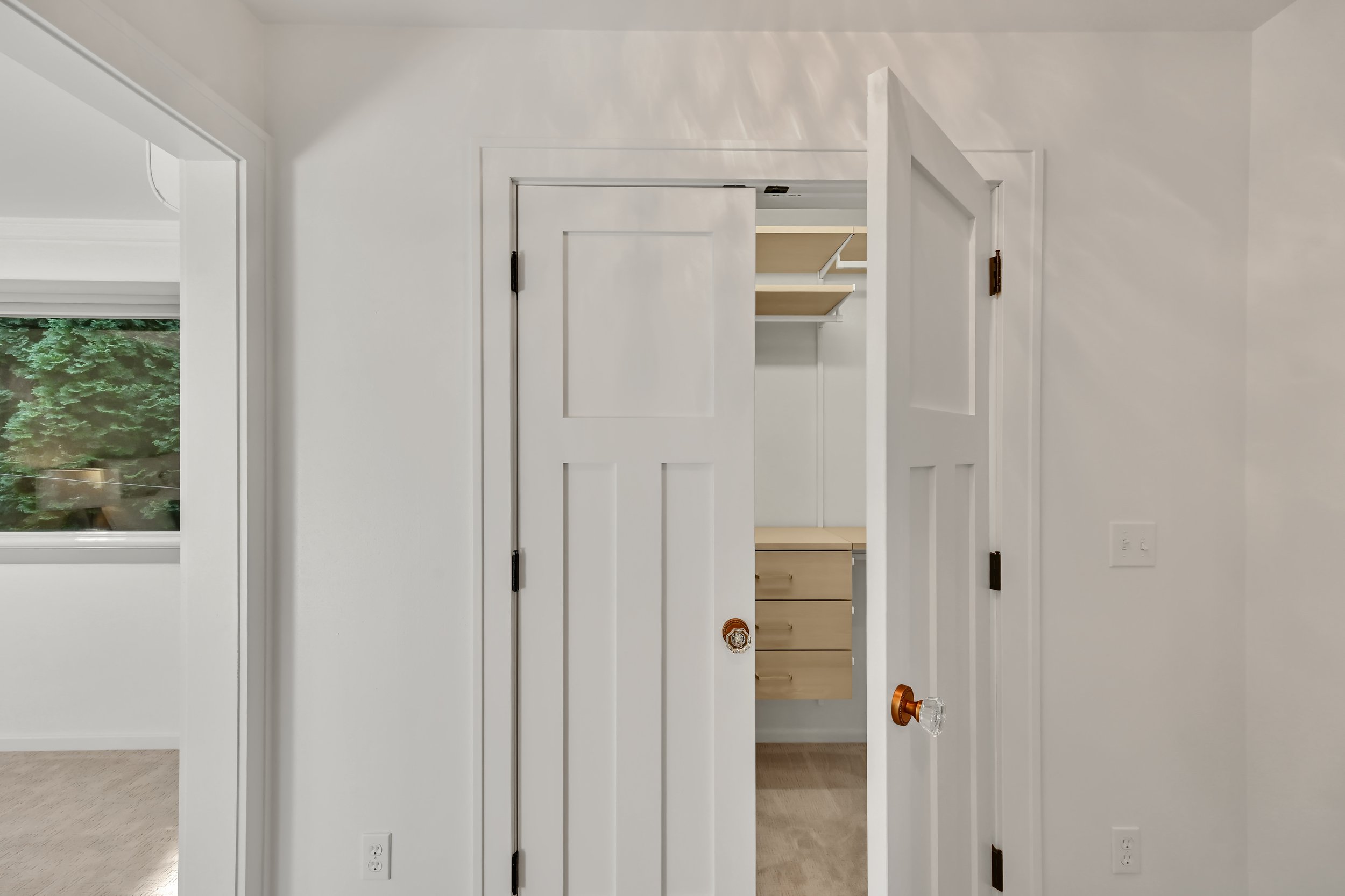 Interior view of a bedroom with white walls, a sliding closet door slightly open showing shelves inside, next to a window with greenery outside, and a copper doorknob on the door.
