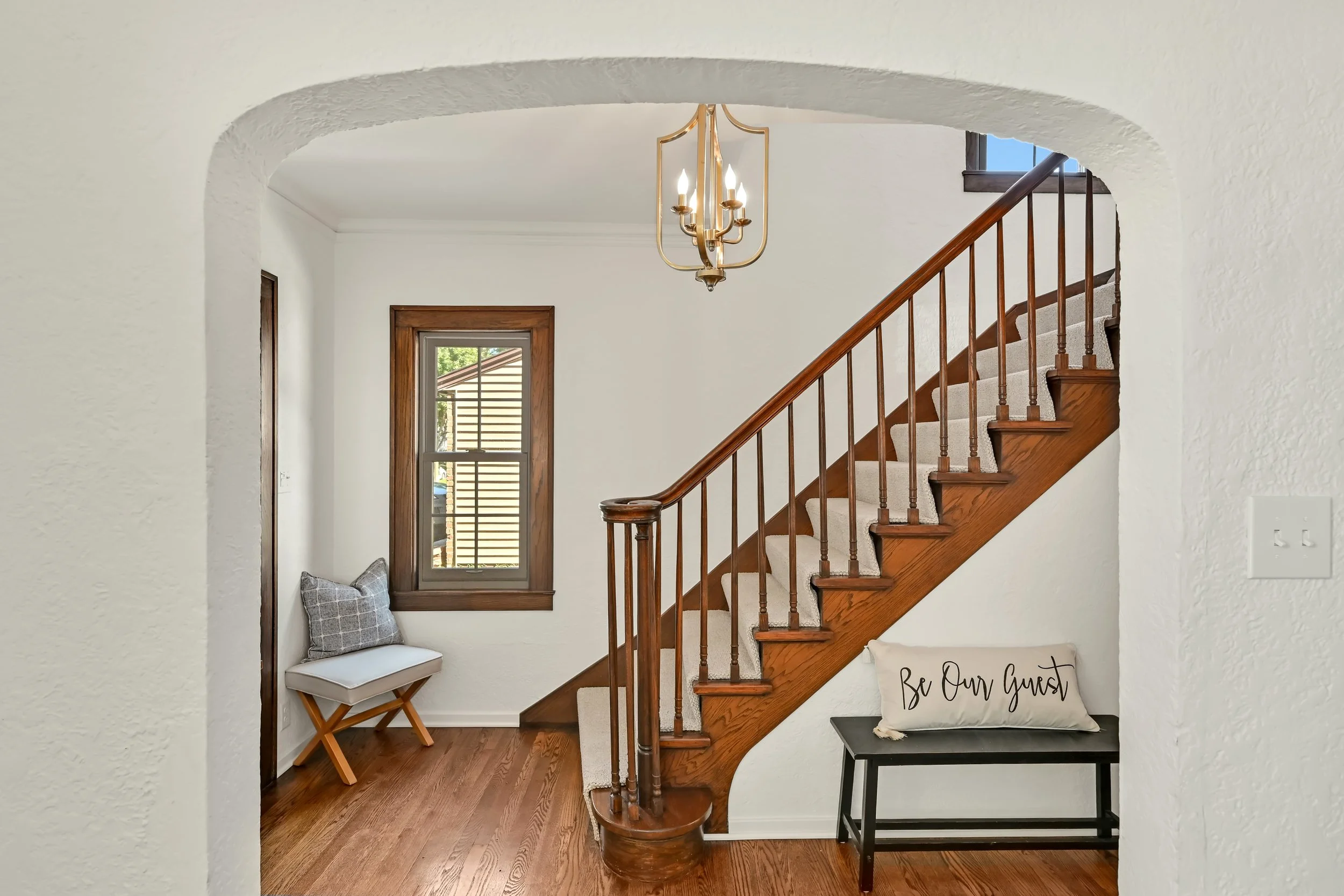 View of a cozy home interior stairway area with hardwood floors and a white textured wall. A small window with wooden trim lets in natural light. A decorative chandelier hangs from the ceiling. There is a wooden stair railing, a small bench or table with a 'Be Our Guest' pillow, and a cushioned seat with a plaid pillow near the window.