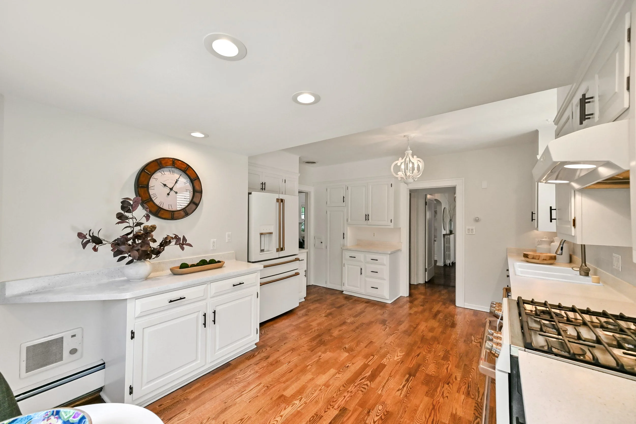 A spacious kitchen with white cabinets, a white countertop, a wall clock, a potted plant on the counter, and a stainless steel stove. There are recessed ceiling lights and a chandelier. The floor is wood, and there are doors and windows in the background.