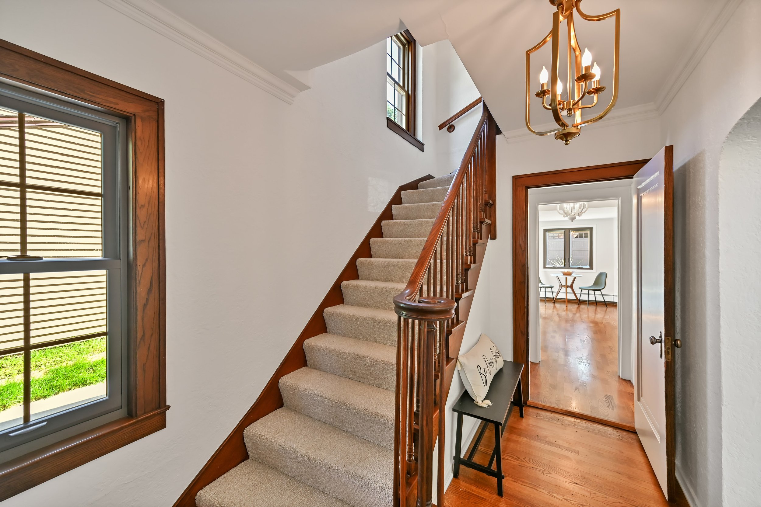 Entryway with carpeted staircase, wooden banister, window with wooden frame, chandelier, doorway leading to a room with hardwood flooring and dining area