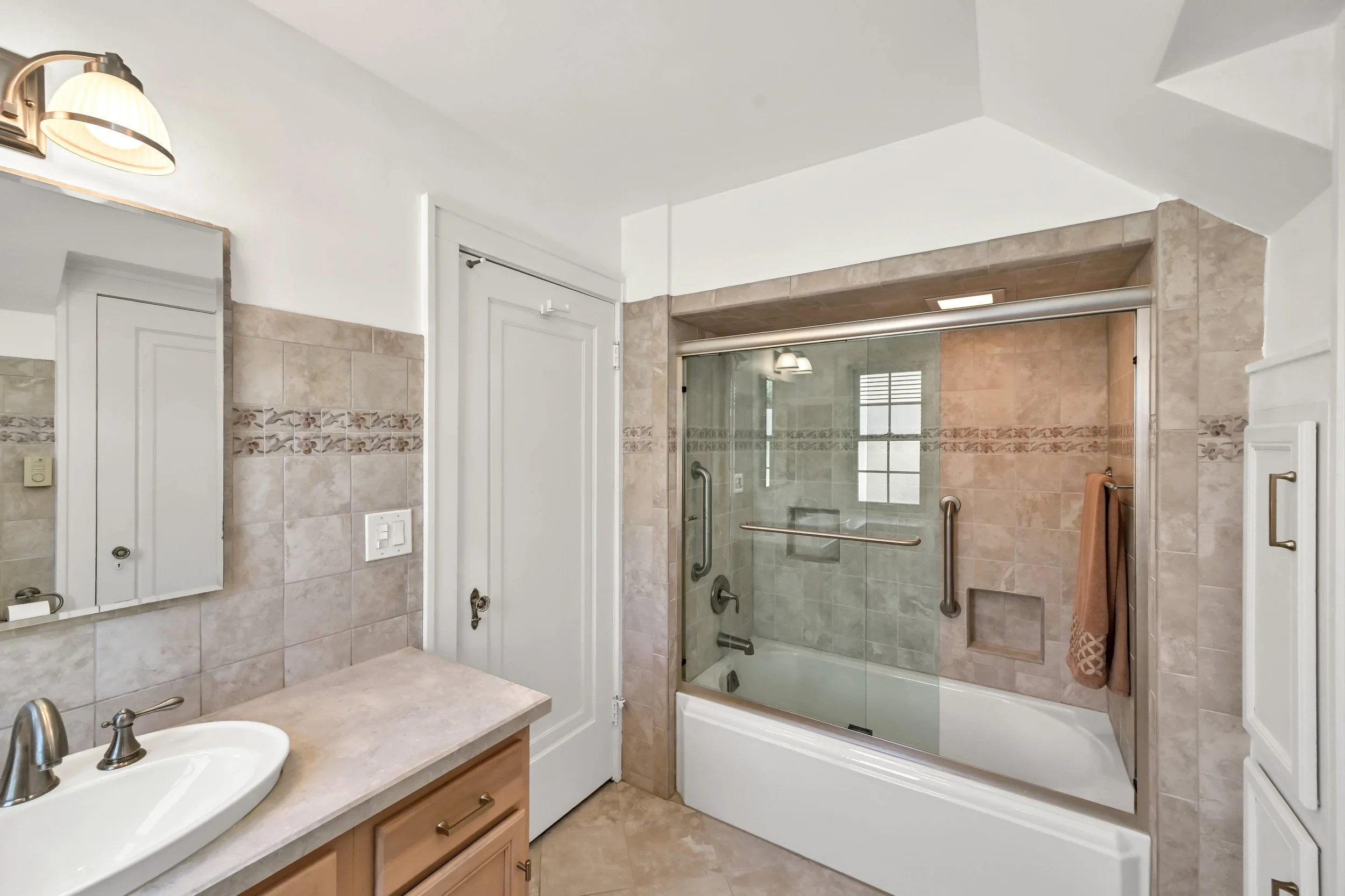 Bathroom with beige tiled walls and floor, a glass shower with a sliding door, a wooden cabinet with a white sink, and a wall-mounted mirror and light fixture.