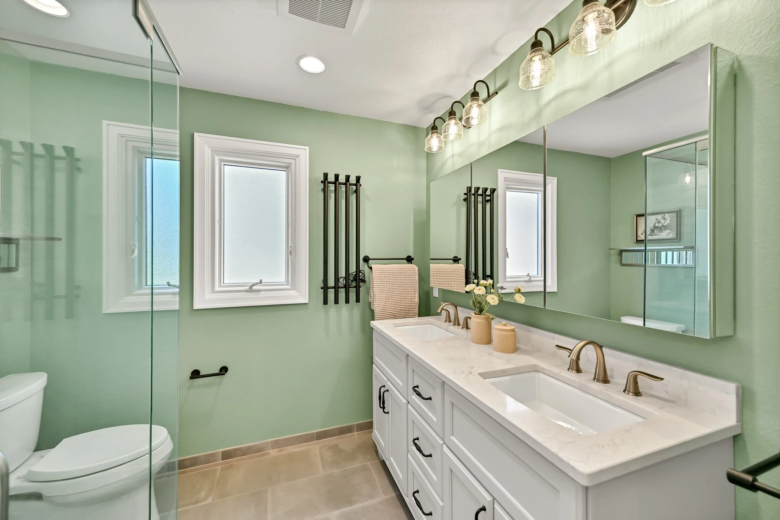 A bathroom with mint green walls, a white vanity with marble countertop, gold fixtures, a large mirror, frosted windows, black towel racks, and a glass shower enclosure.