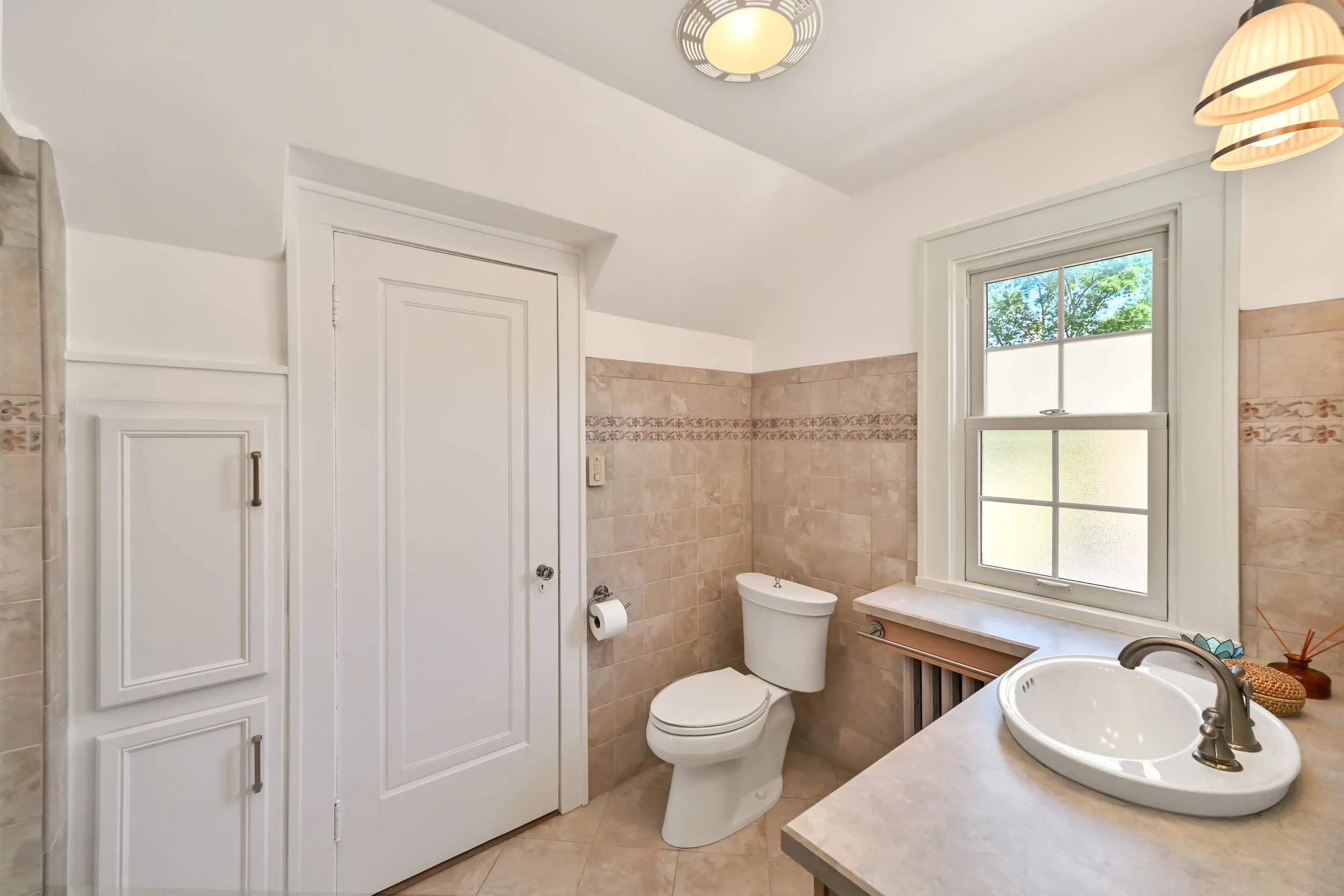 A bathroom with beige tiled walls and a white toilet next to a window with a view of green trees. There is a white cabinet with a handle, a round sink with a faucet on a beige countertop, and a vanity with a wicker basket and decorative items. The ceiling has a round light fixture and two pendant lights with lampshades.