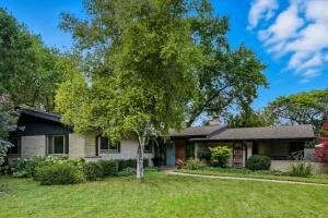A single-story house with a large front yard, green lawn, and trees, under a blue sky with some clouds.