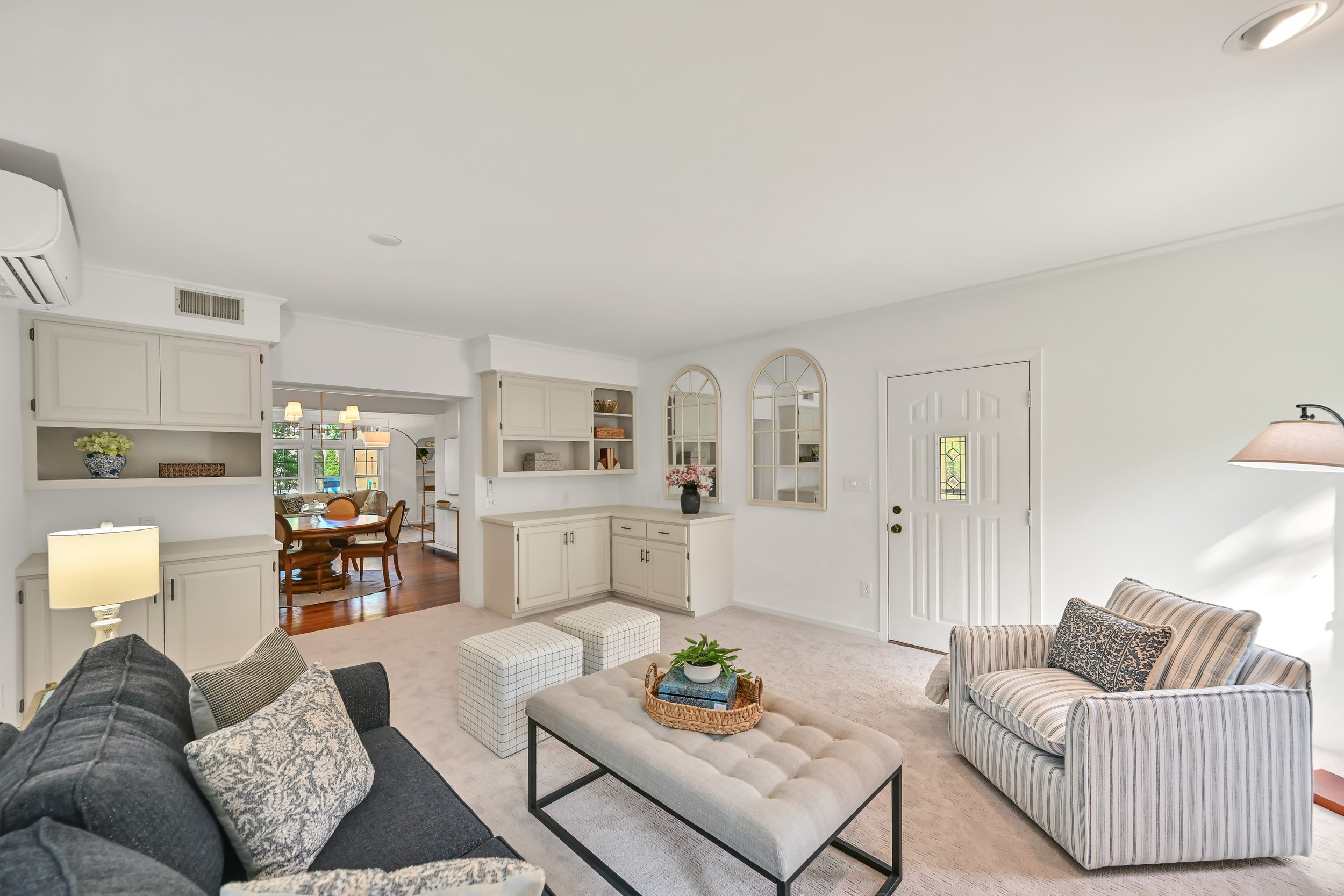 Living room with white walls, beige carpet, black sofa with patterned pillows, striped armchair, coffee table with plant, opening to dining area with wood furniture, built-in white shelves, arched mirrors, and front door visible.