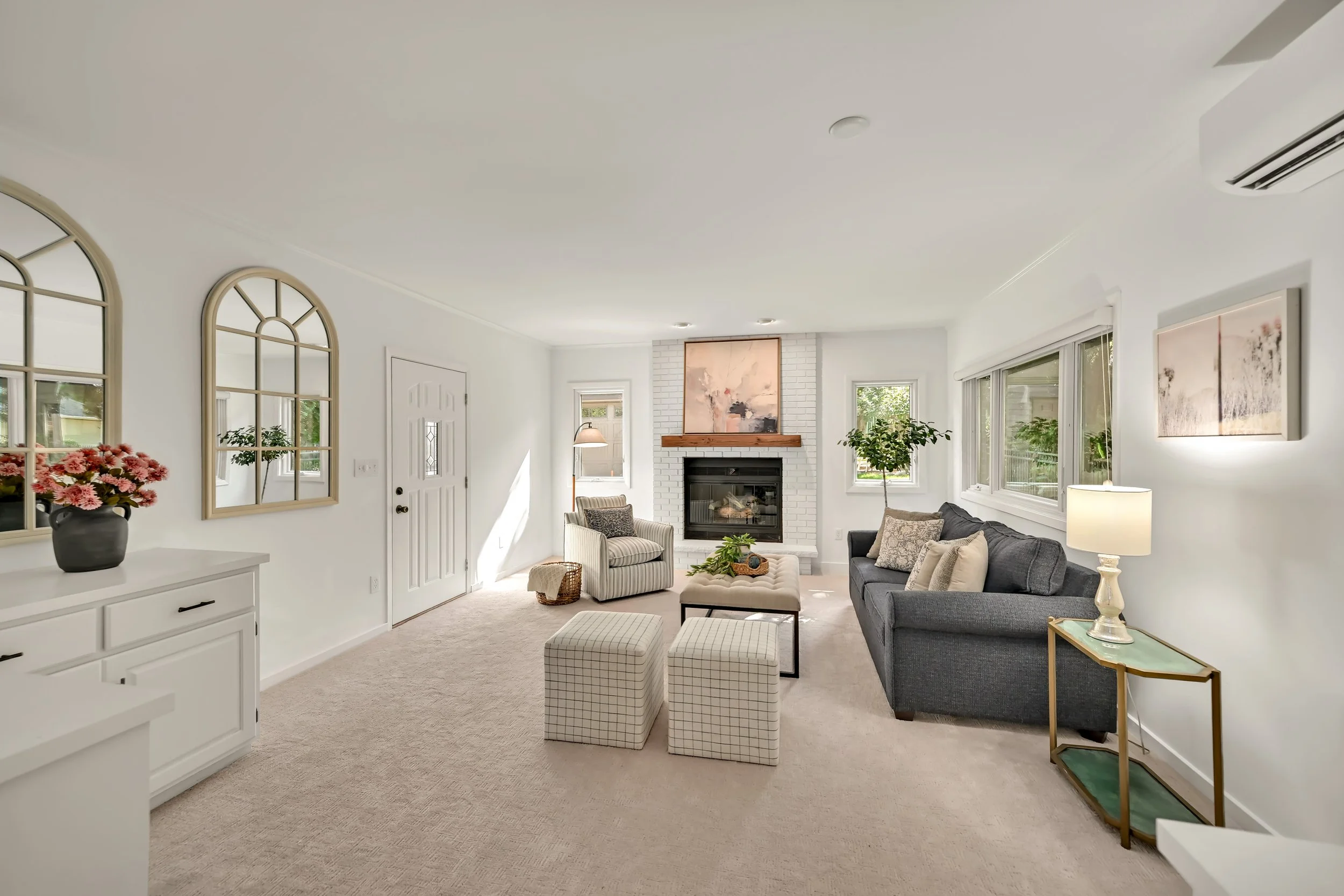 Bright living room with white walls, fireplace, and large windows, decorated with plants, artwork, and furniture including a gray sofa and striped armchair.