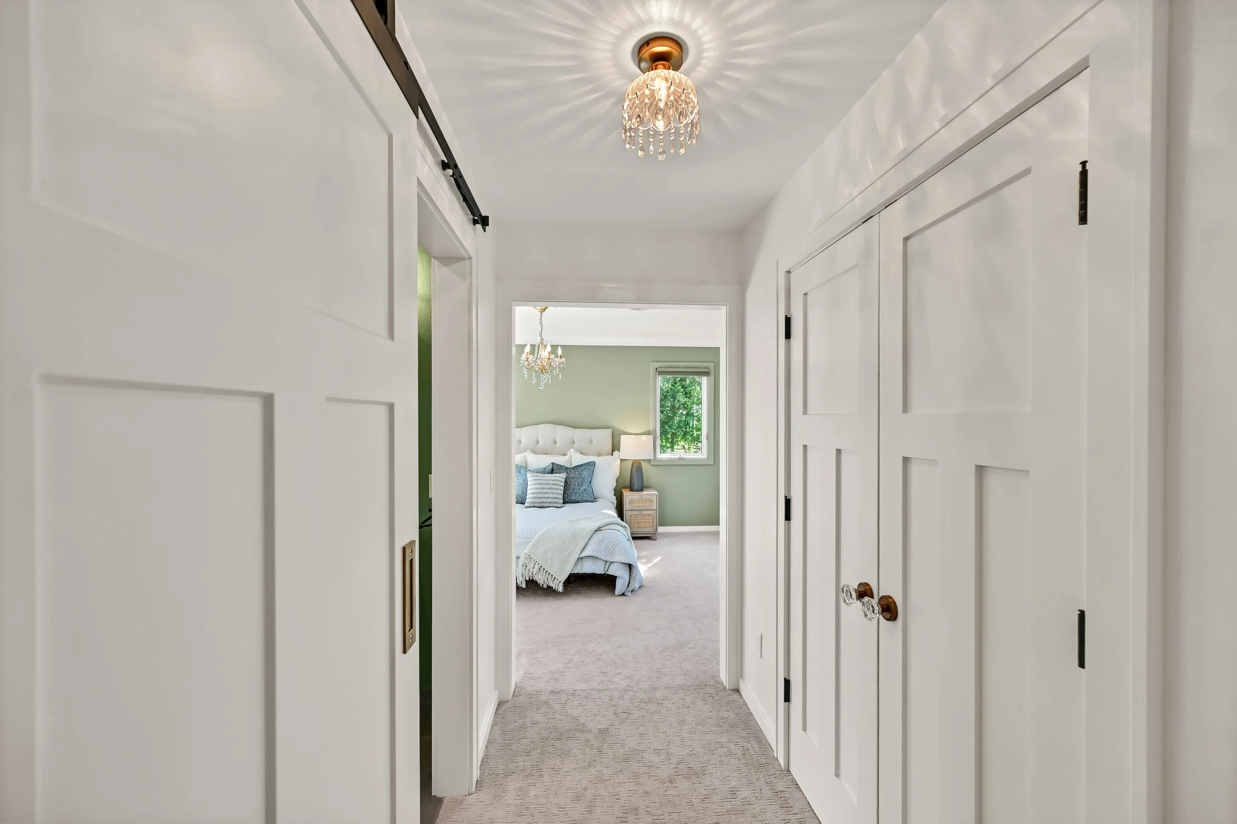 A view down a hallway with white walls and closet doors, leading to a bedroom with a bed, pillows, a lamp, and a window with greenery outside, illuminated by a chandelier.