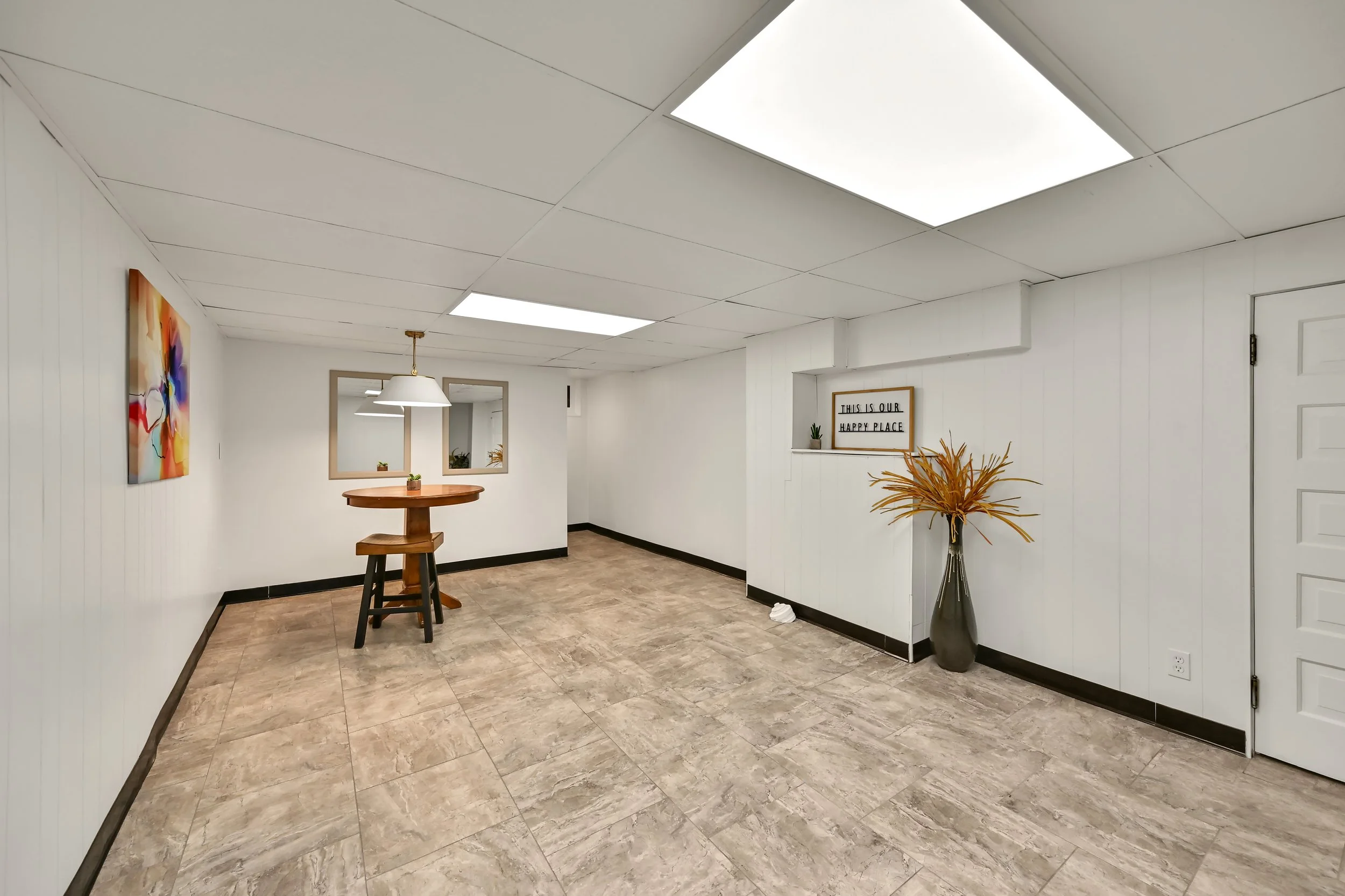 Interior of a room with white ceiling and walls, brown tiled floor, a wooden table with a stool, abstract painting on the wall, mirrors, framed sign that reads 'This is our happy place,' a large vase with dried plants, and ceiling fluorescent lights.