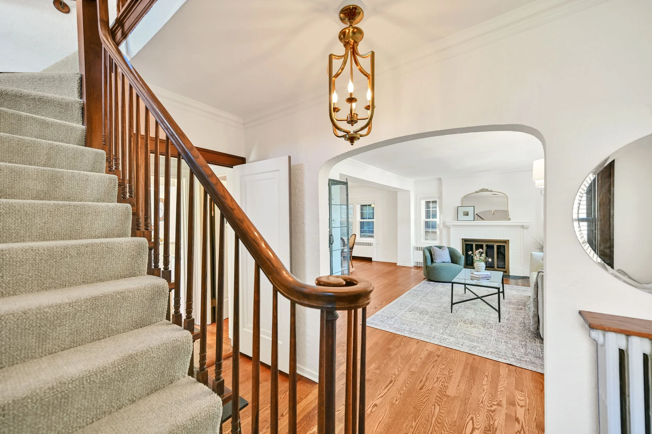 View from the staircase looking into a living room with a white fireplace, mirror, and seating area, illuminated by a gold chandelier. Whitefish Bay tudor.