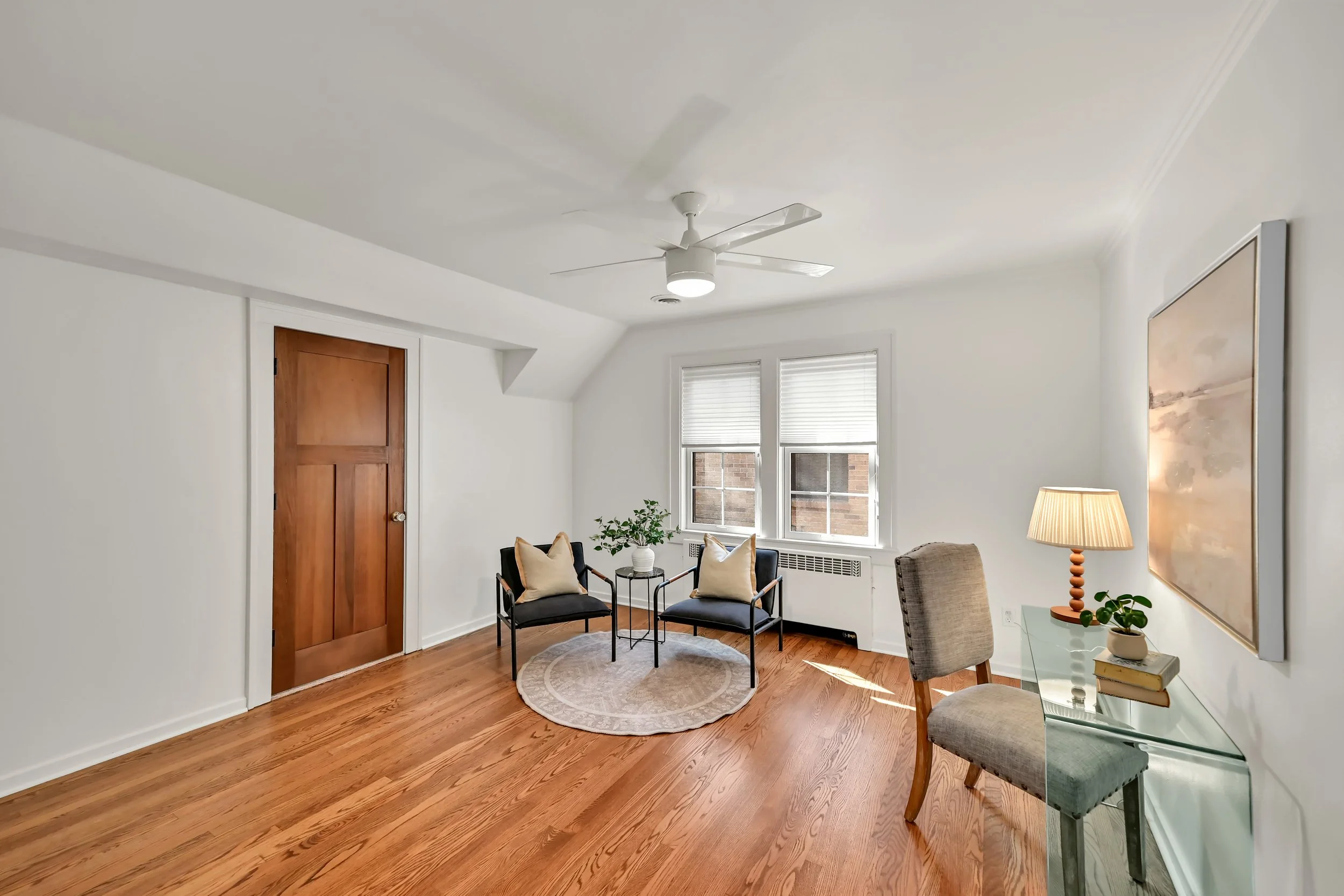 A bright living room with hardwood floors, white walls, and a ceiling fan. There are two black chairs with beige pillows, a small round rug, a wooden door, three windows with blinds, a table with a lamp, and a framed artwork on the wall.