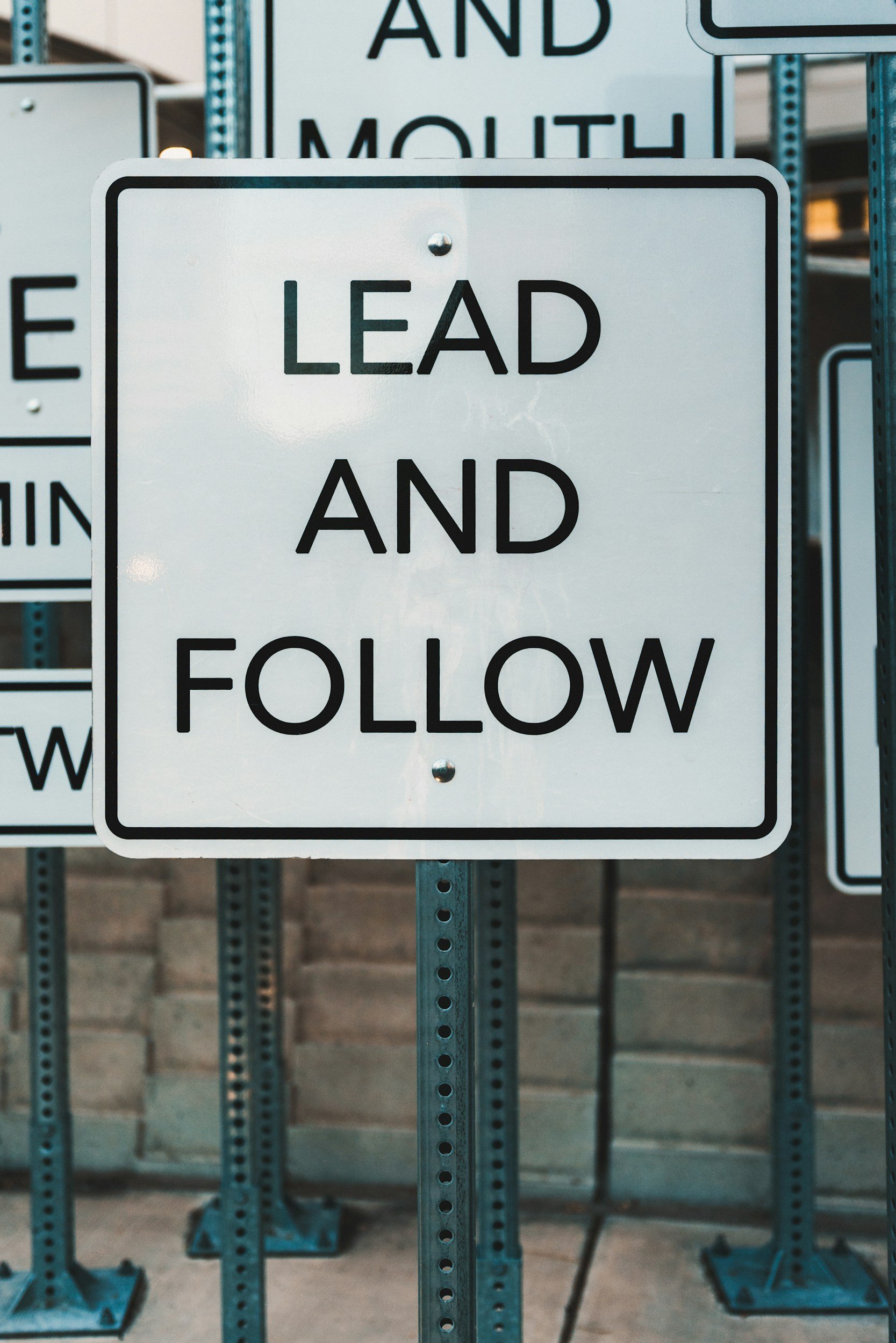 Close-up of a white road sign reading “LEAD AND FOLLOW,” surrounded by other street signs. Learn more about joining our team at uliyan.com