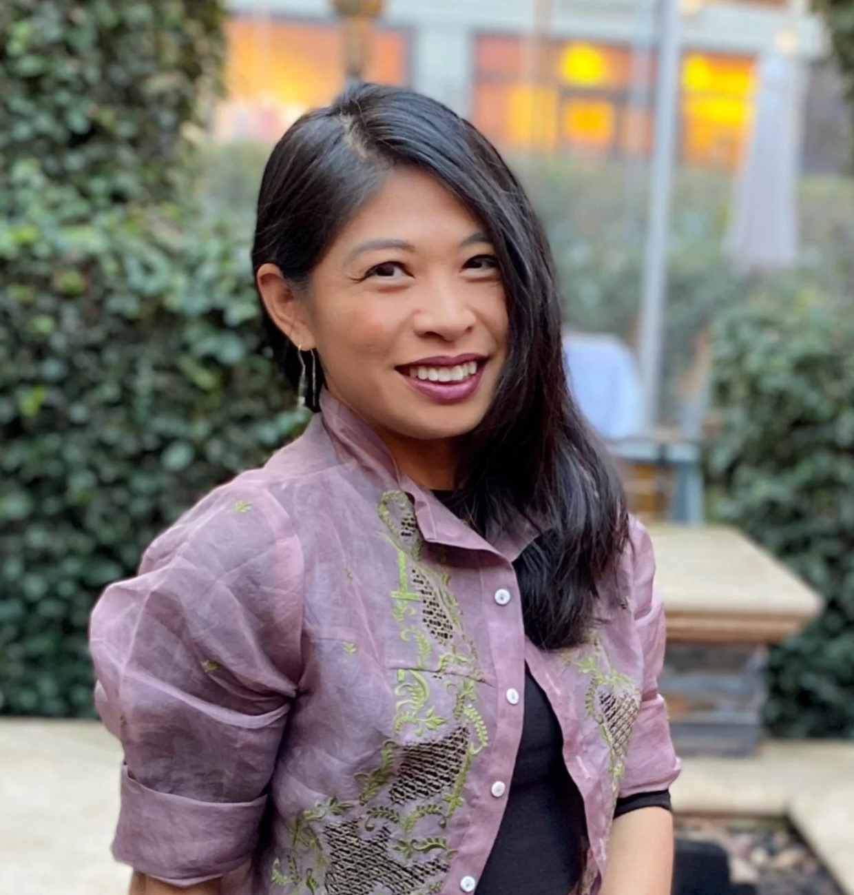 A pinay with long black hair and earrings smiling outdoors in front of green bushes, wearing a pinkish-purple embroidered sarong Tagalog blouse.