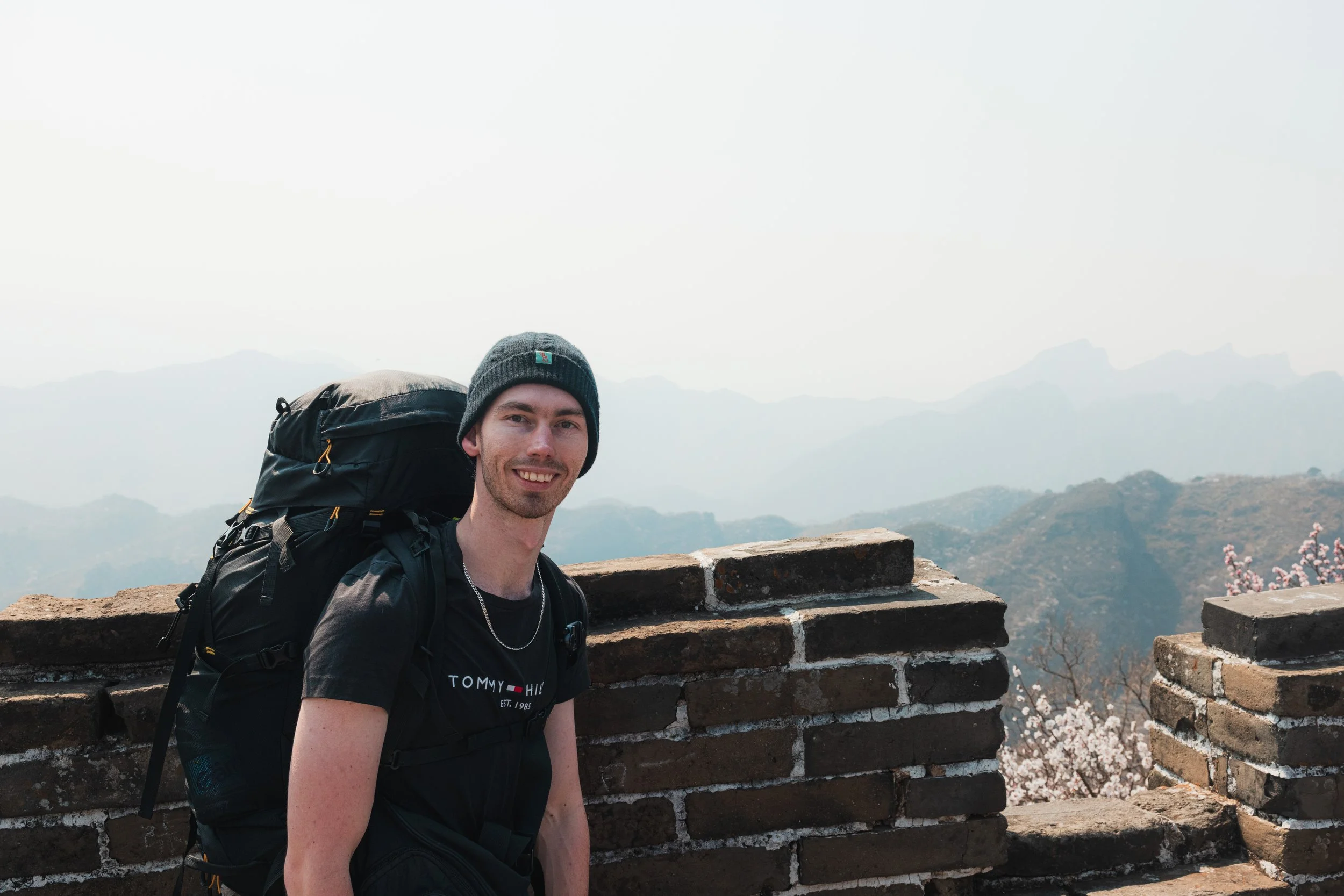 A smiling man with a backpack standing at a historic site with a brick wall, mountains, and trees in the background, wearing a gray beanie and a black t-shirt, during daytime.