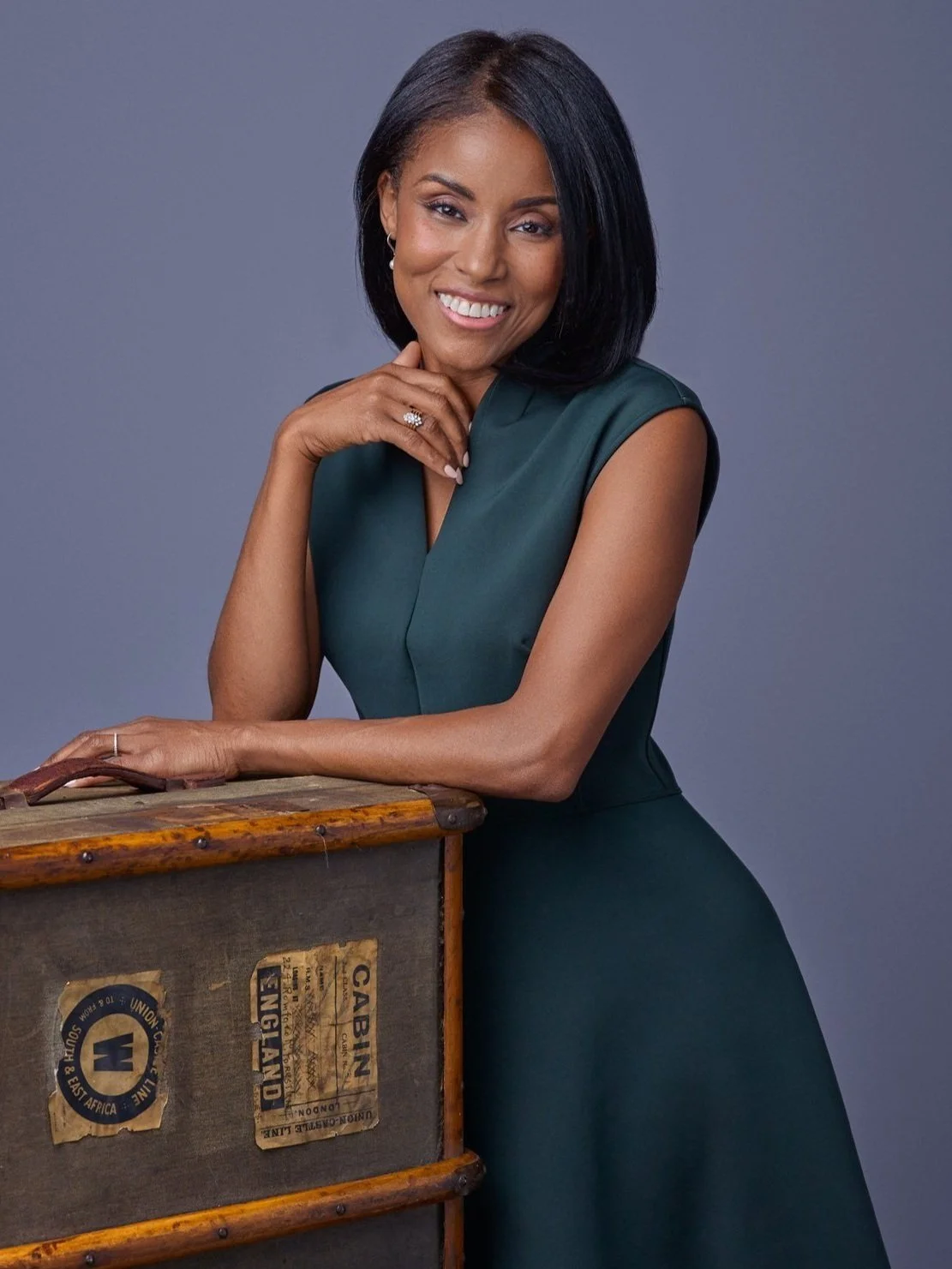 A woman with medium skin tone and shoulder-length black hair smiling with her hand near her chin, wearing a dark green sleeveless dress, standing next to a vintage wooden trunk against a plain gray background.