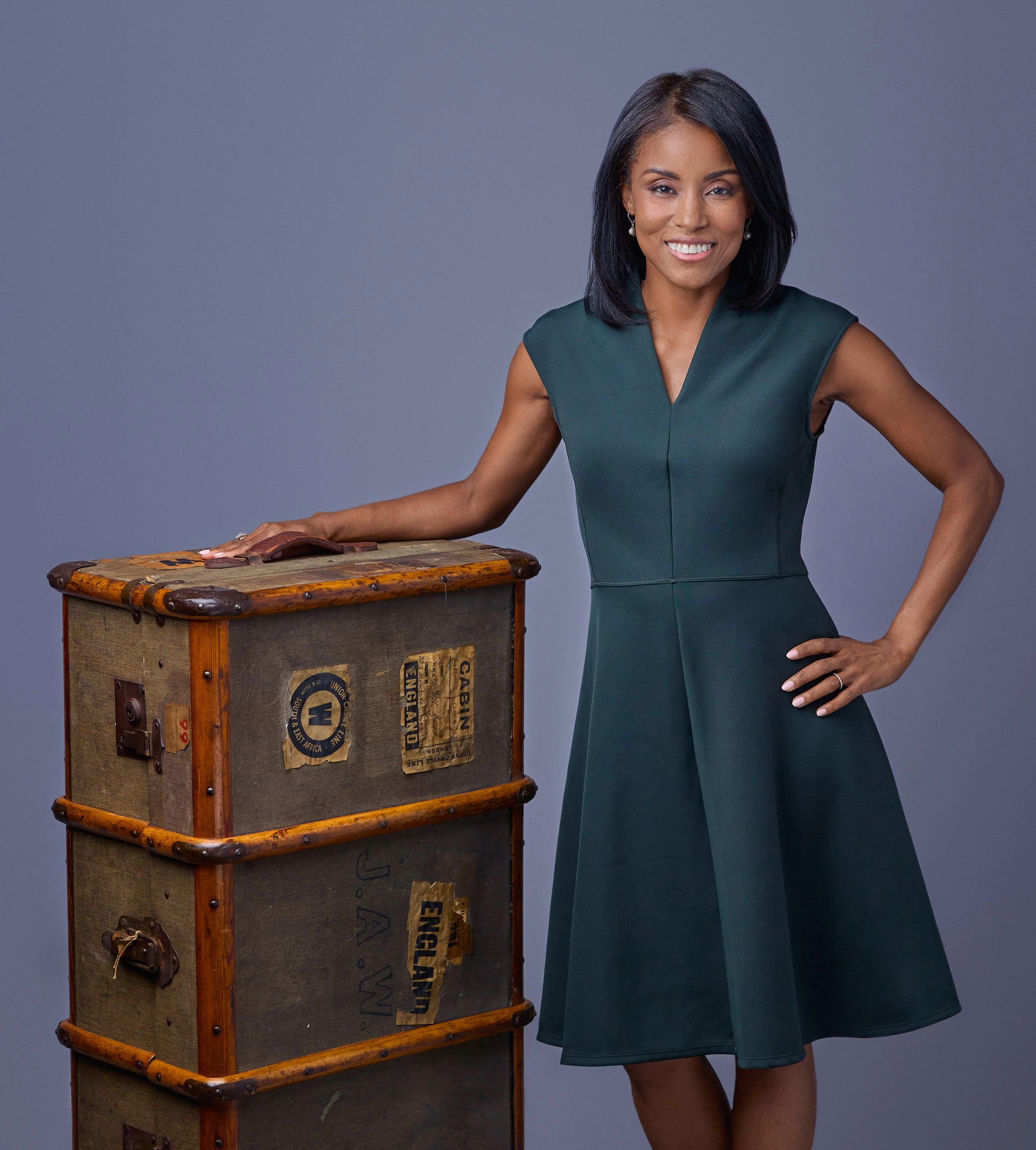 Woman with black hair, wearing a dark green sleeveless dress, standing next to an old vintage wooden suitcase with stickers and labels, posing with her right hand on her hip against a gray background.
