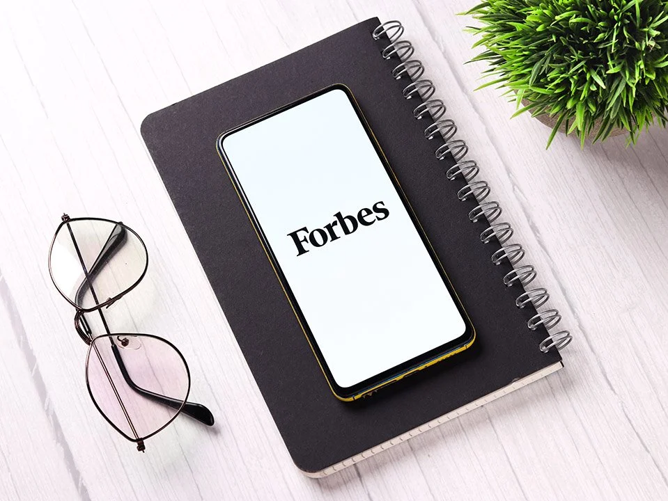 Smartphone displaying Forbes logo, placed on a black spiral notebook, with eyeglasses, a small green plant, and a white wooden desk surface.