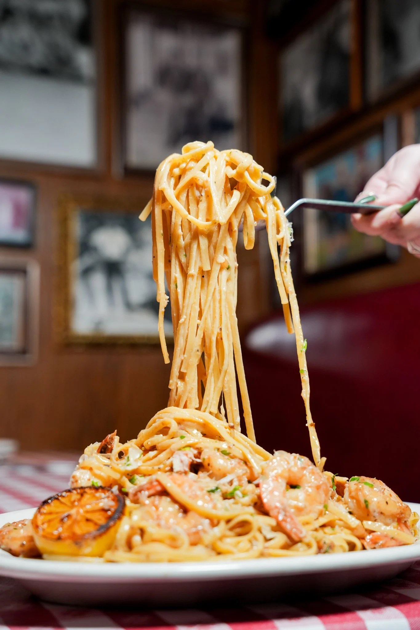 A plate of shrimp and pasta, with a close-up of cooked spaghetti being lifted by a fork. The pasta appears to be in a creamy sauce with lemon slices and herbs, set on a table with a red and white checkered tablecloth. Background shows a wooden wall with framed pictures.