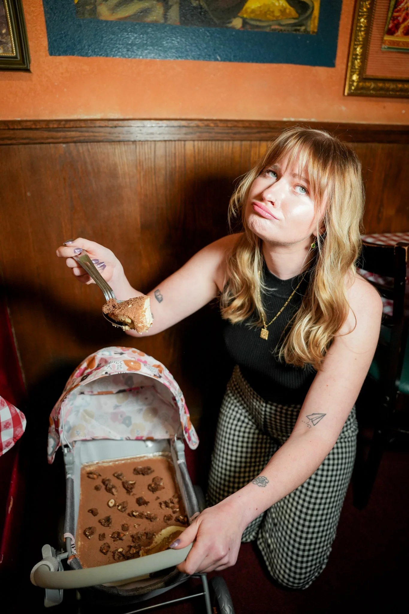 A woman with blonde hair, tattoos, and wearing a black top and checkered pants is sitting in a restaurant, holding a fork with a piece of chocolate cake. There is a stroller in front of her with the remaining cake on it.
