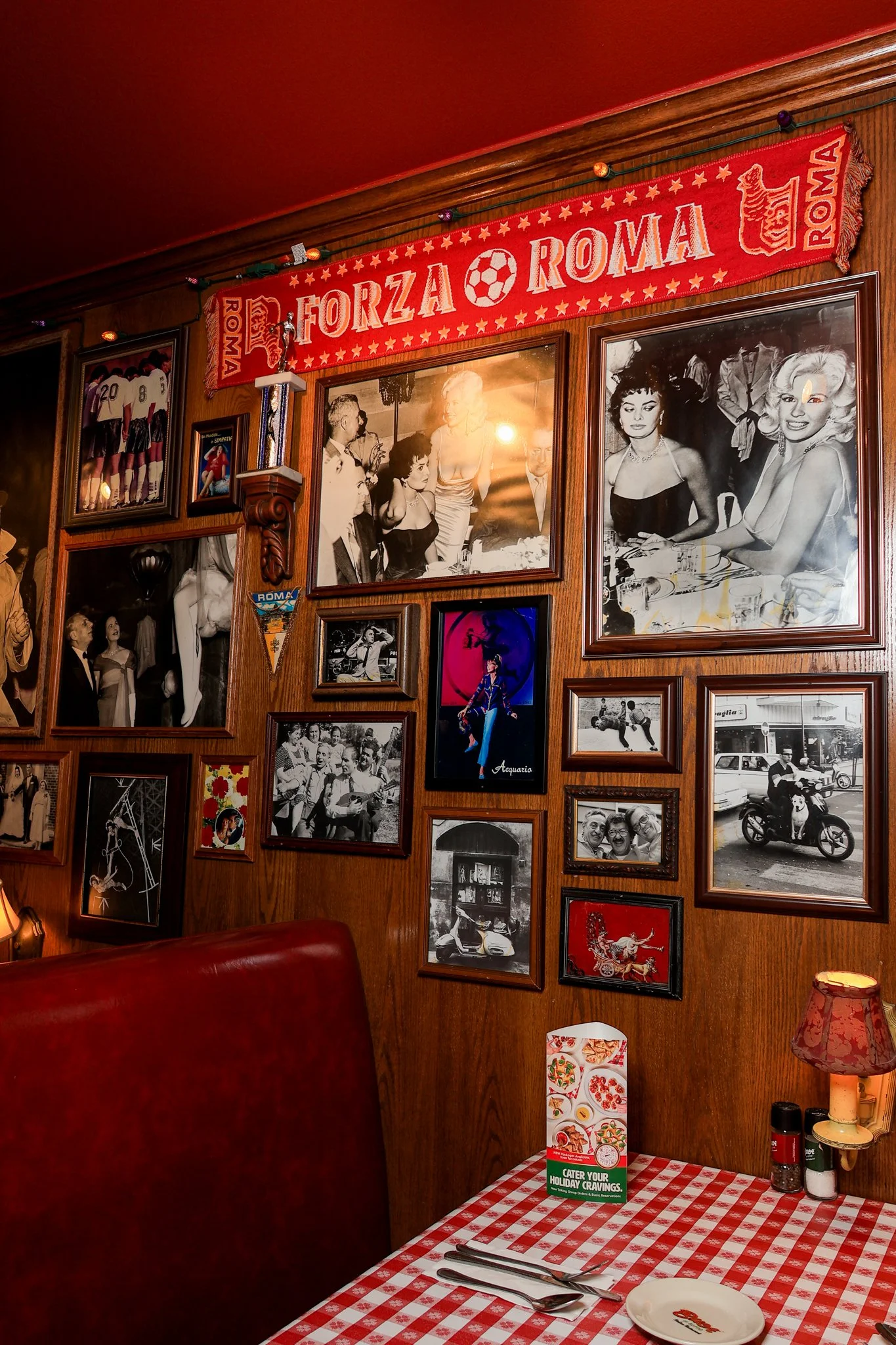 Wall decorated with framed photos and memorabilia, a red and white checkered tablecloth on a table with plates, utensils, and condiments, a small lamp, and a red and white scarf reading "Forza Roma" hanging above.