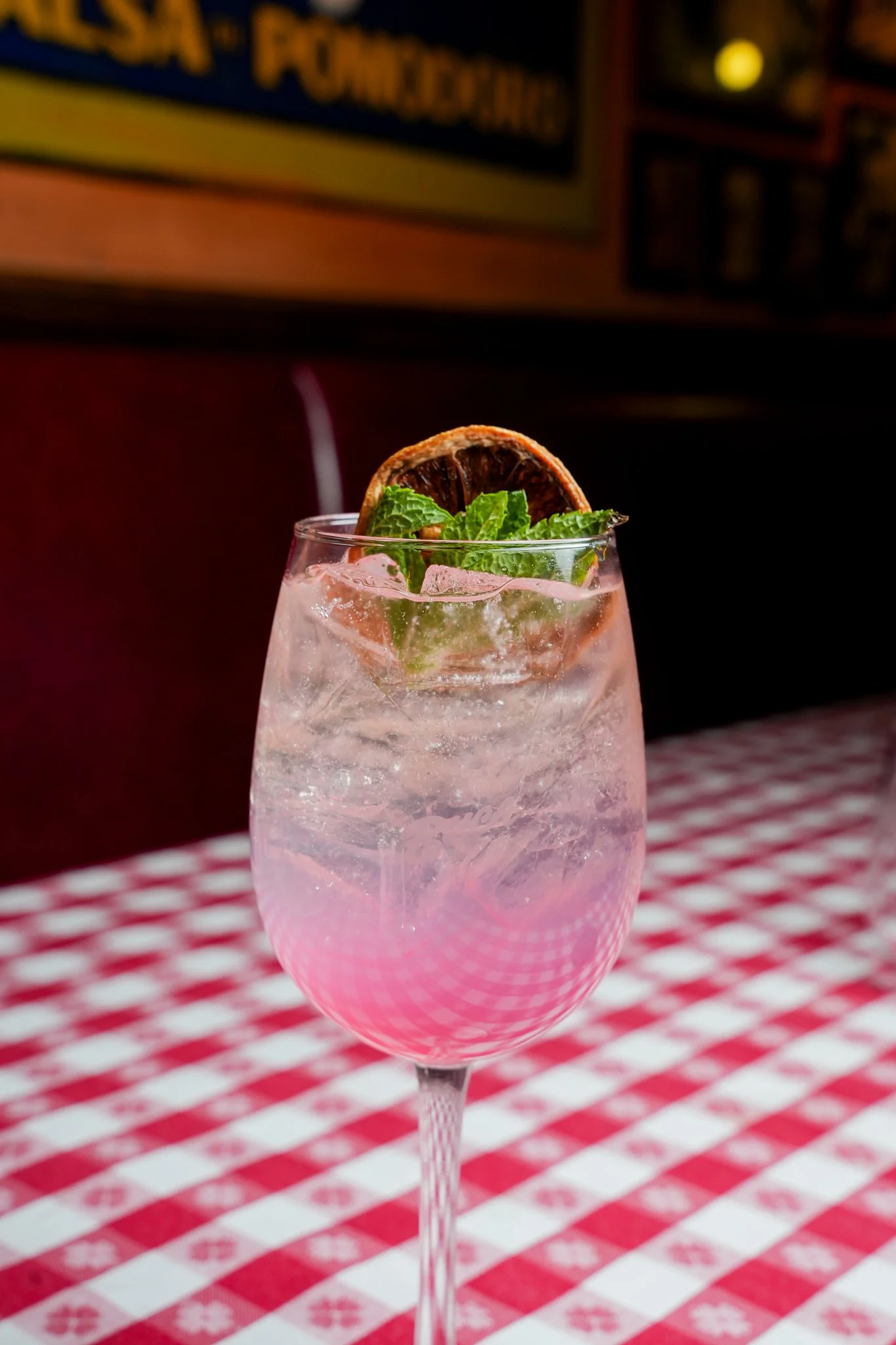 Pink cocktail in a wine glass garnished with a dried lime slice and mint leaves on a red and white checkered tablecloth.
