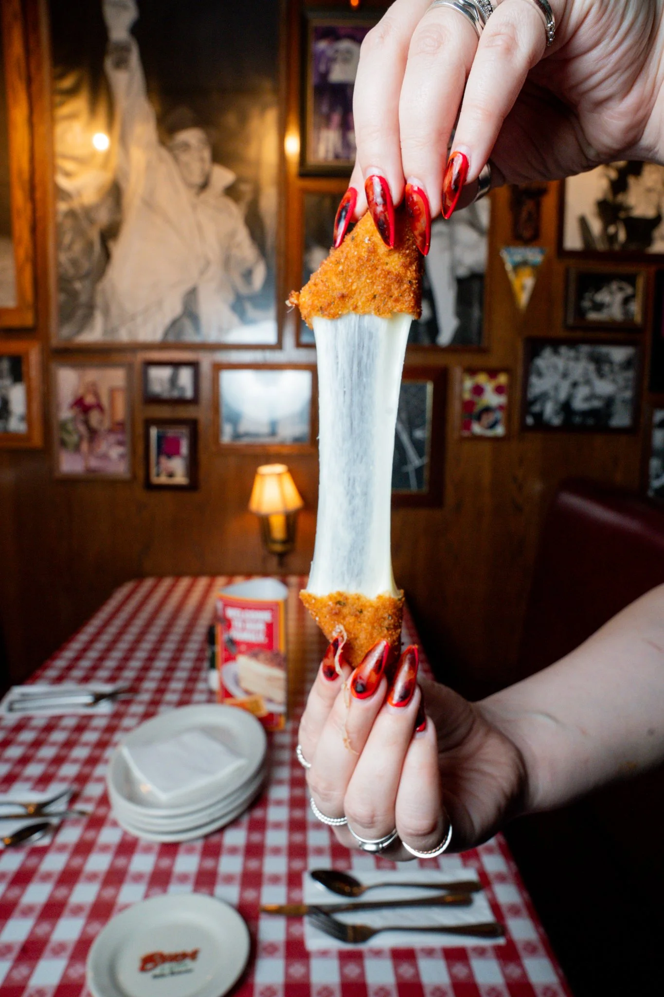 Person with painted nails holding a cheese bun with cheese stretching in the middle at a restaurant with a red checkered tablecloth and framed photos on the wall.