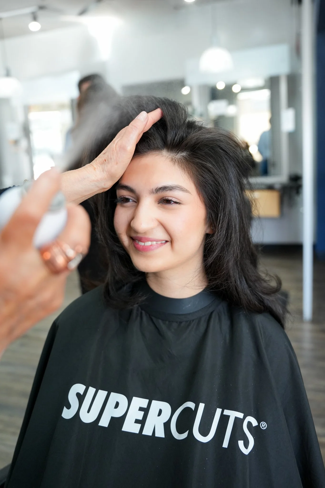 Young woman at a hair salon getting her hair styled, wearing a black cape with white text 'SUPERCUTS'.