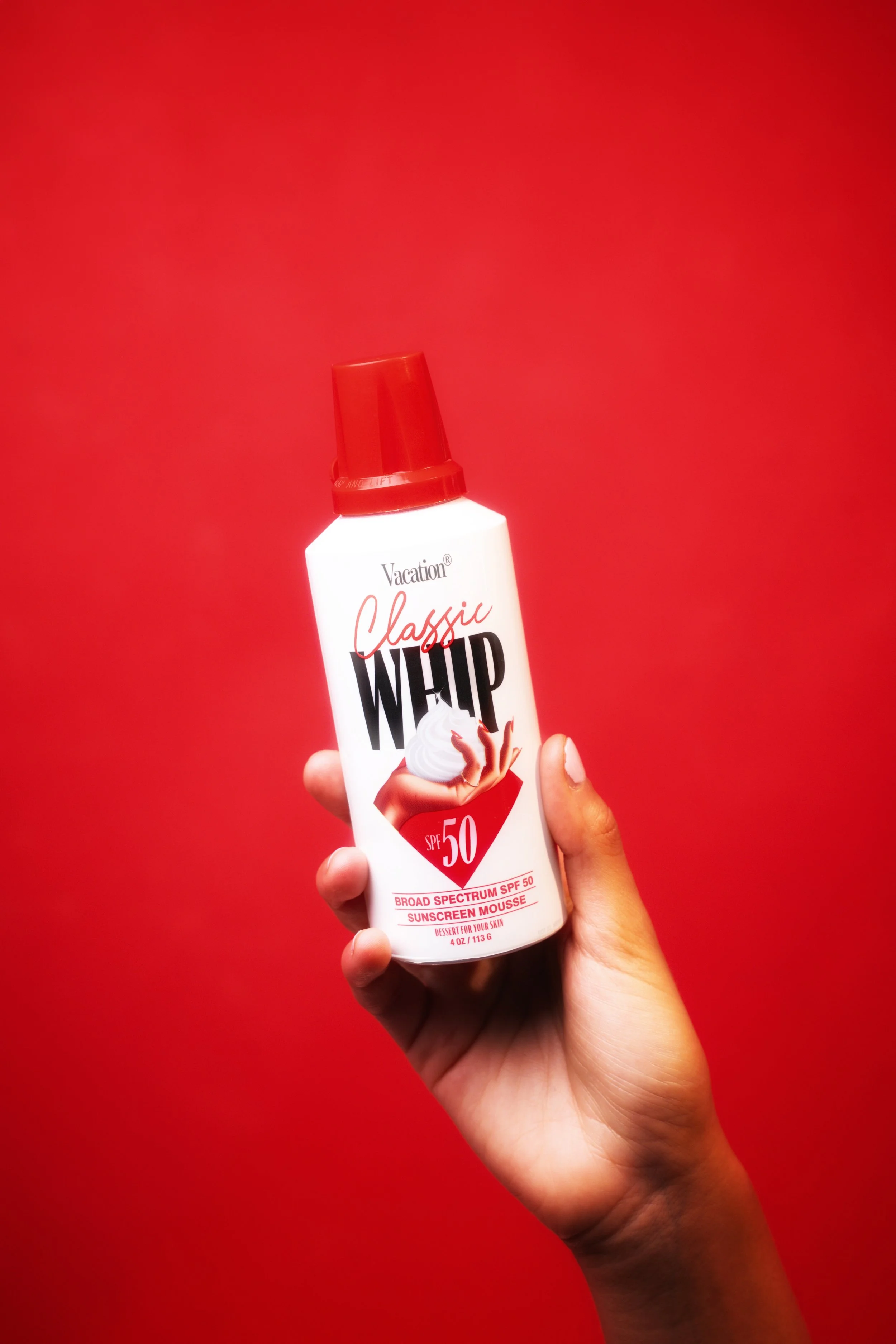 Hand holding a bottle of Vacation Classic Whip Sunscreen Mousse SPF 50 against a red background.