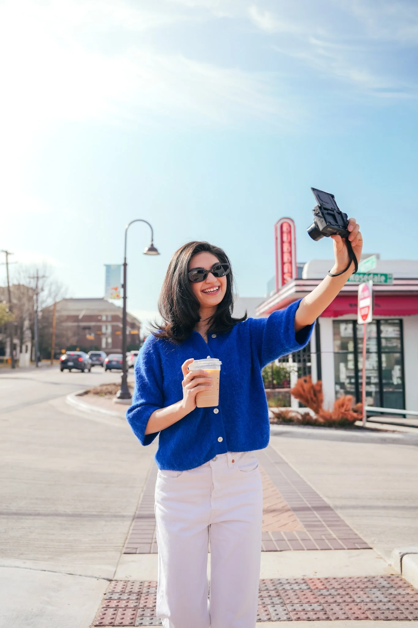 Young woman with black hair, sunglasses, and a bright blue sweater, smiling and taking a photo with a camera on a street with a coffee in hand.