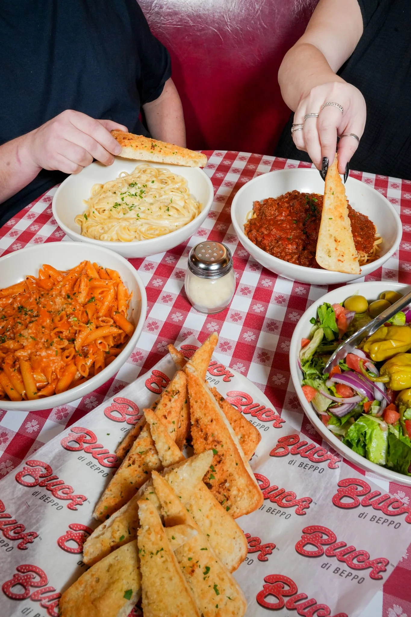 A table with various Italian dishes including pasta, salad, garlic bread, and a bowl of pasta with meat sauce, with two people sharing the meal.