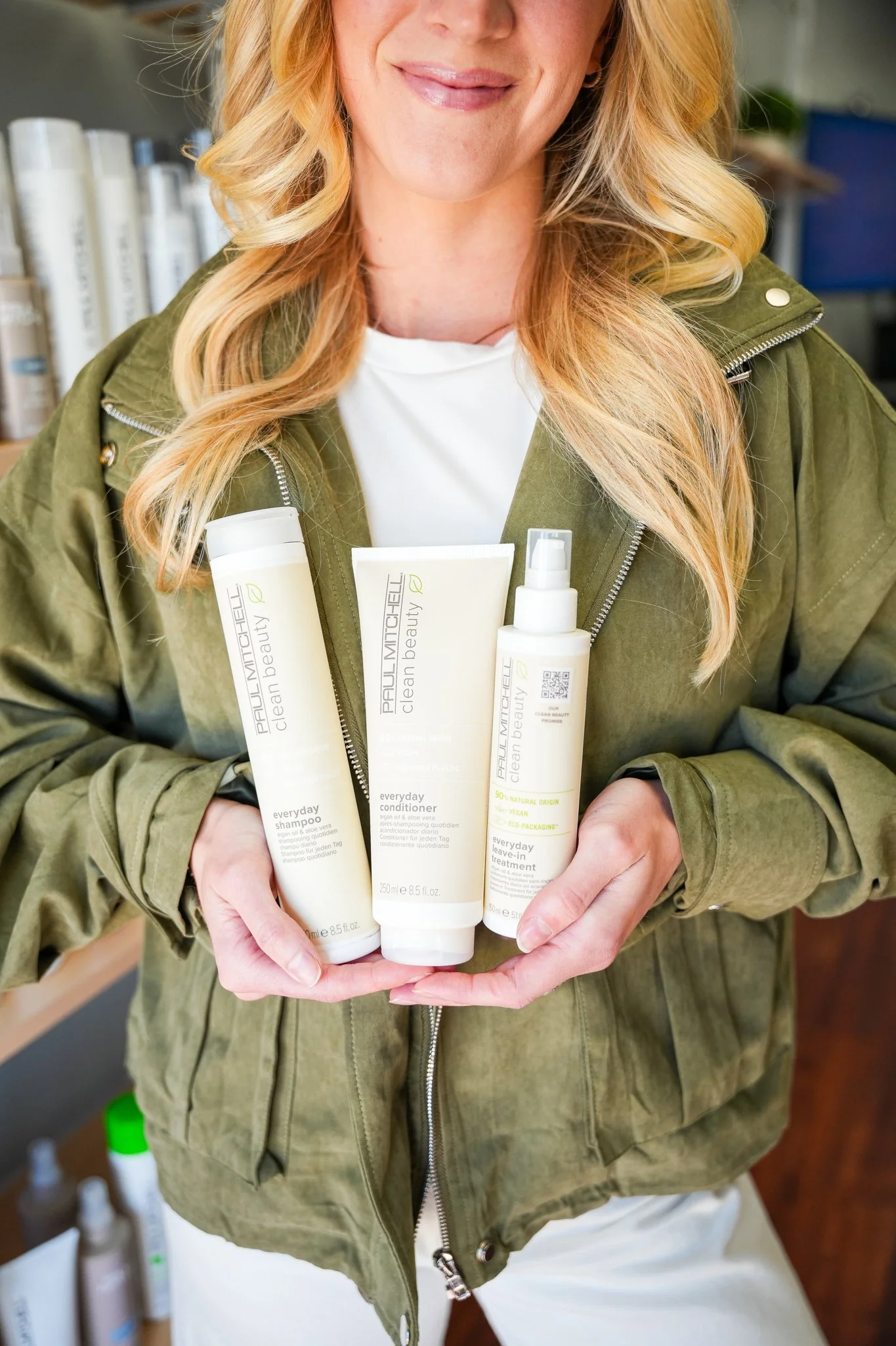 A woman holding three Paul Mitchell Clean Beauty everyday hair care products, including shampoo, conditioner, and leave-in treatment, in a store or salon setting.
