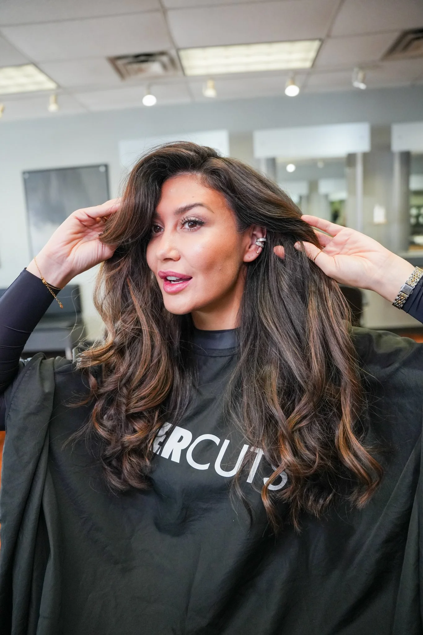 A woman with long, wavy brown hair, wearing a black salon cape with the word 'CIRCUITS' partially visible, is getting her hair styled in a salon.